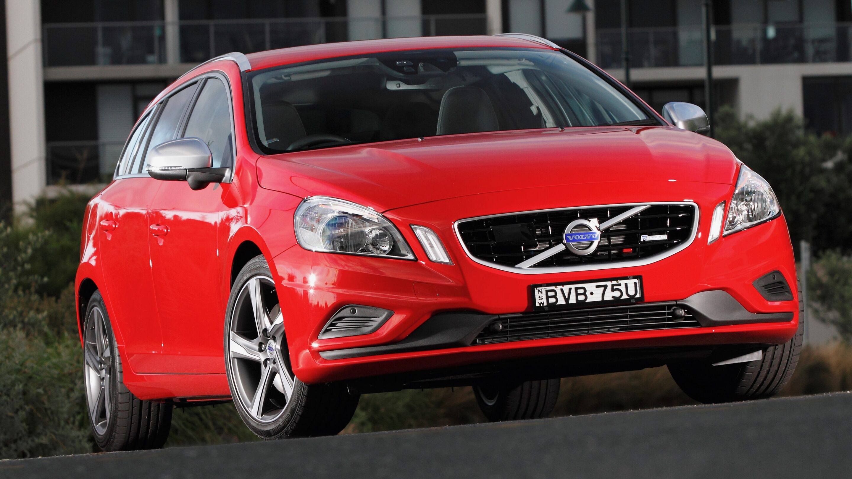 Front-right angled view of a bright red Volvo SUV showcasing its grille and headlights, with an urban building backdrop and visible license plate BVB-75U.