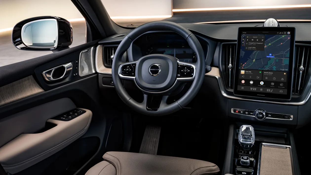 Interior of a Volvo car: steering wheel, large center touchscreen, gear selector, and beige leather seats seen from the driver’s side.