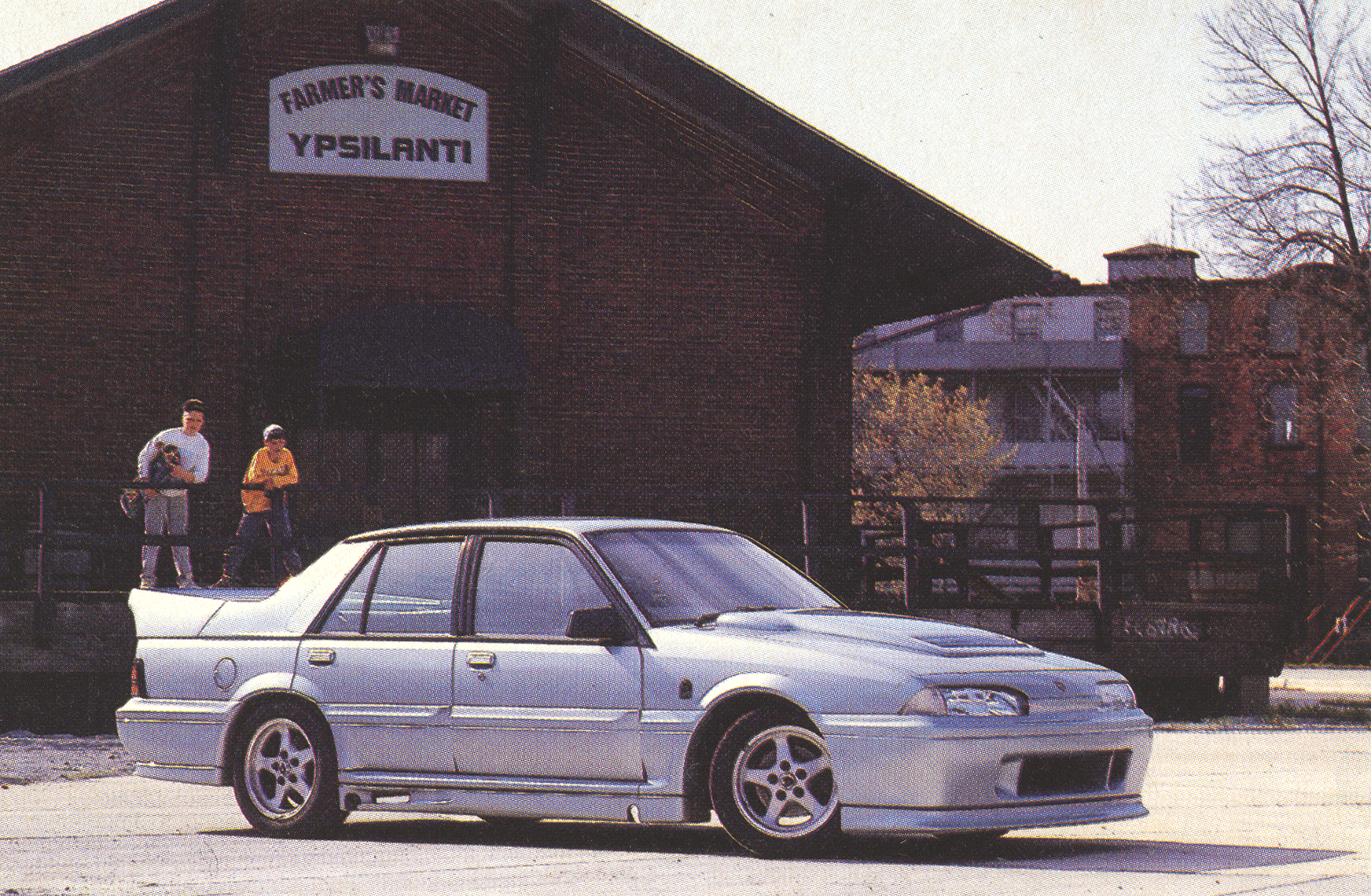 Silver four-door sedan parked in a lot in front of a brick Farmers Market building with a sign reading 'FARMER'S MARKET YPSILANTI'; three people stand near the left side of the building.