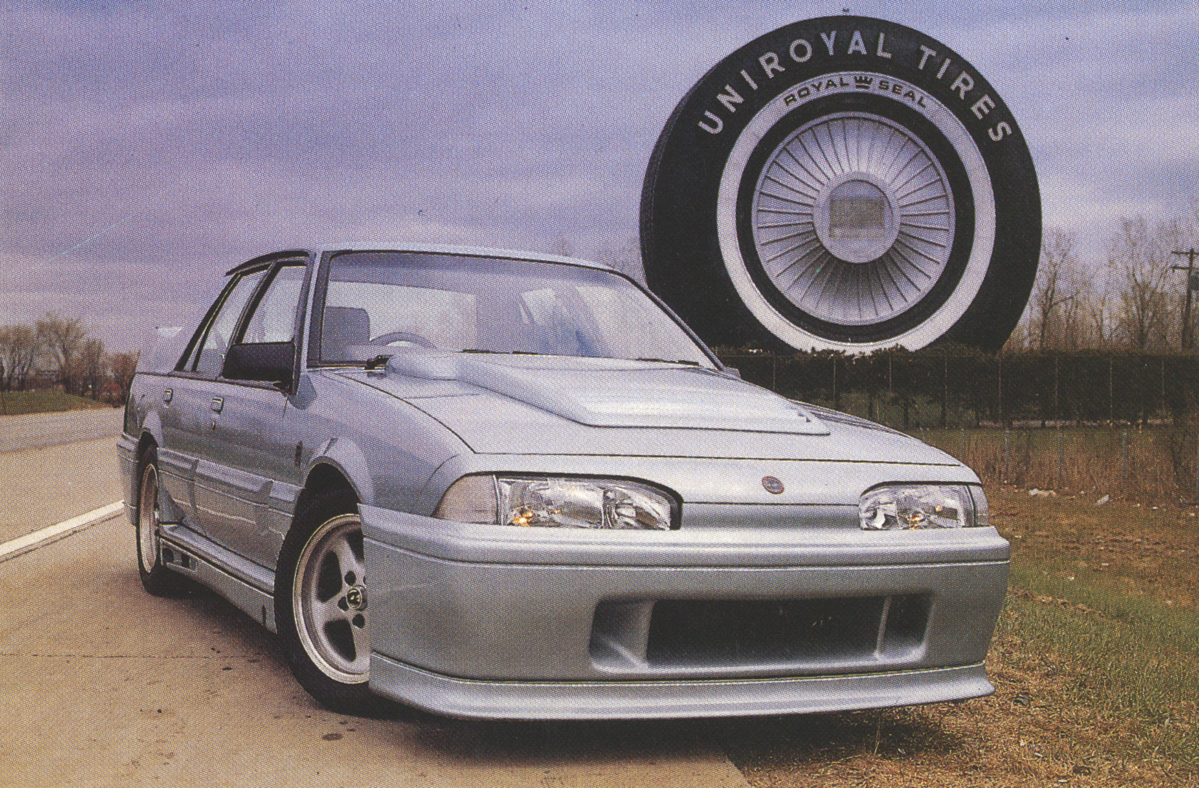 Silver sedan foreground on a dirt road with a giant tire sculpture reading UNIROYAL TIRES in the background.