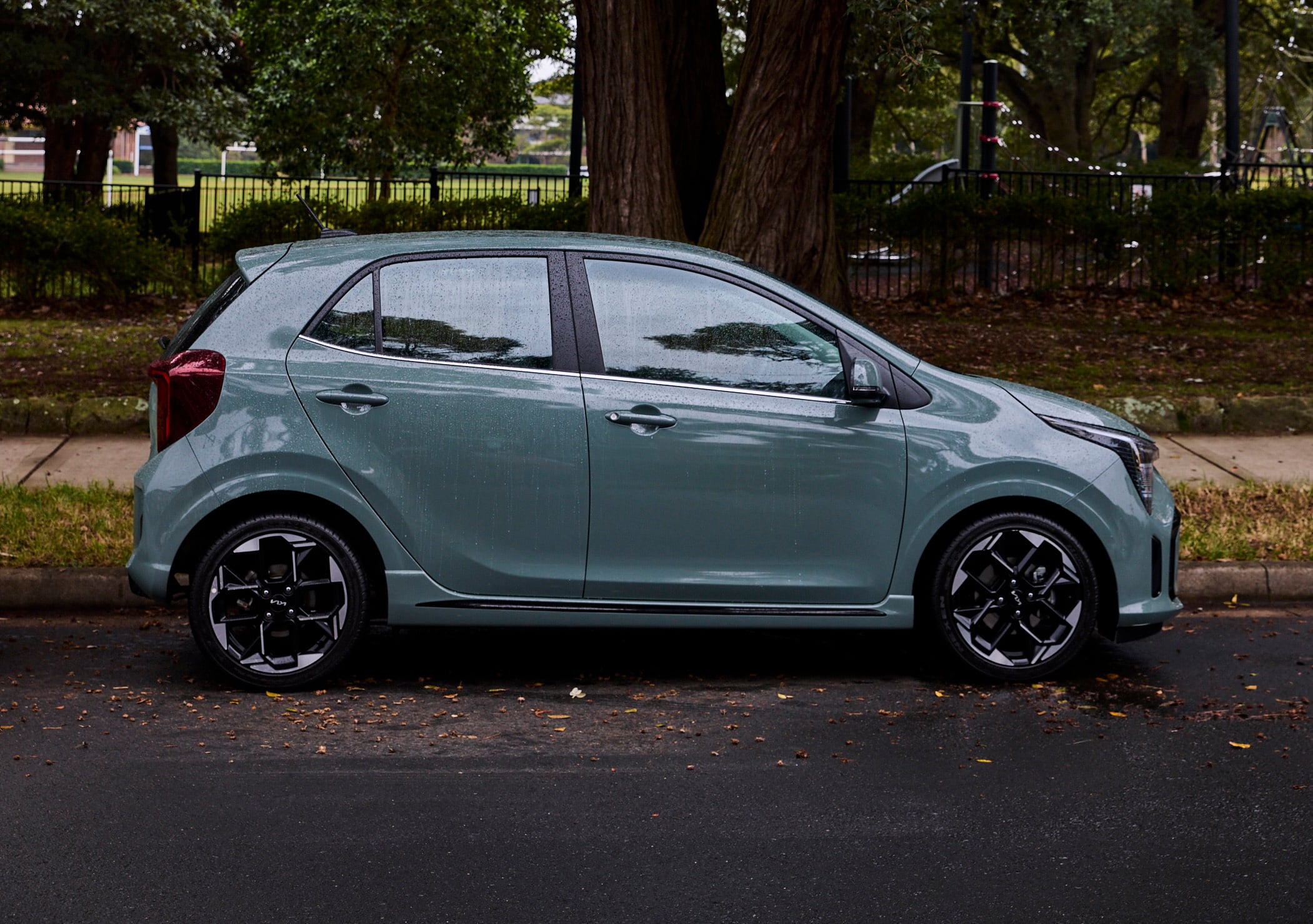 Side view of a teal compact car parked on a wet street with rain droplets on the windows and a park with trees in the background.