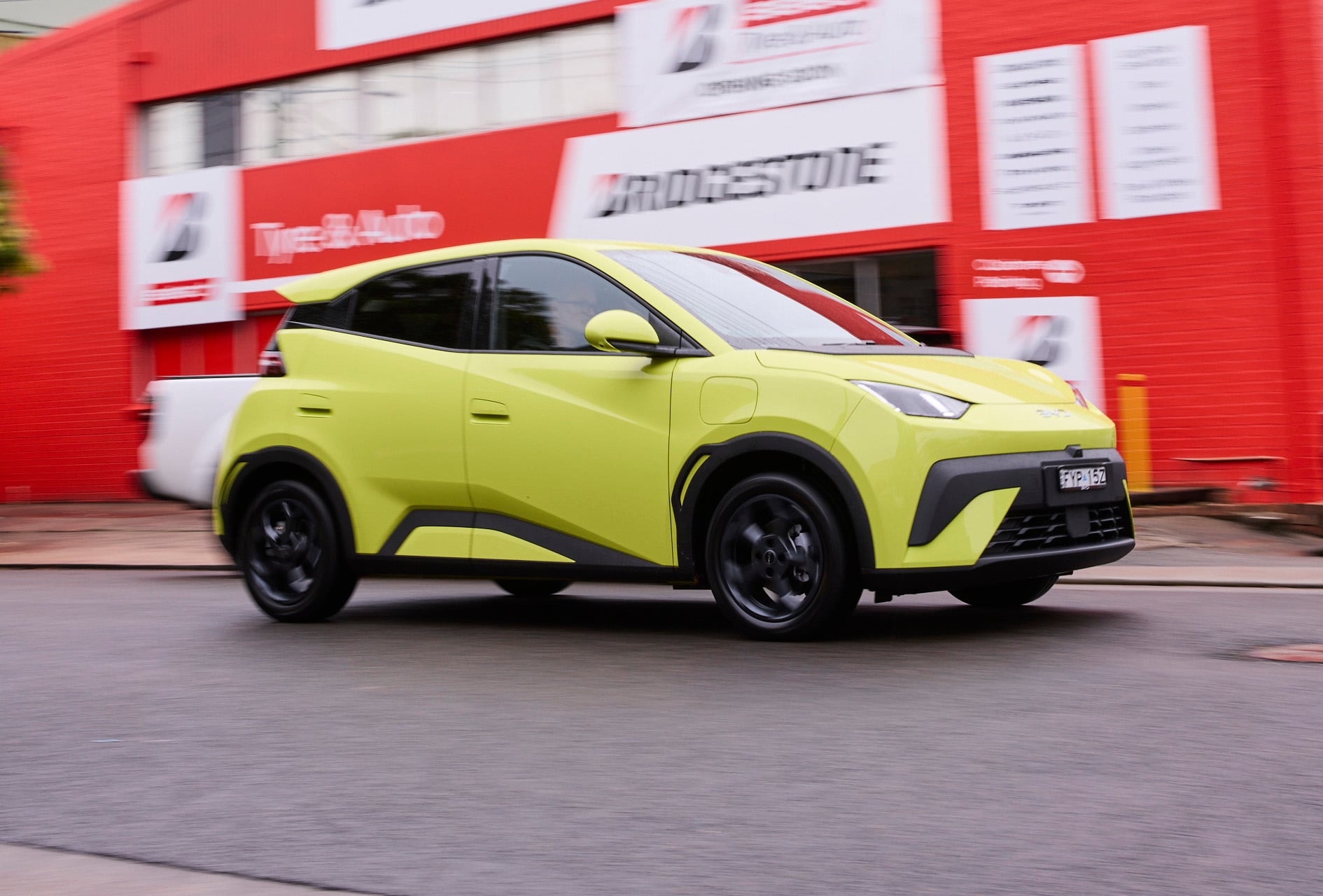 Bright lime-green SUV speeding by a red auto shop building with blurred signage in the background, motion blur emphasizes speed.