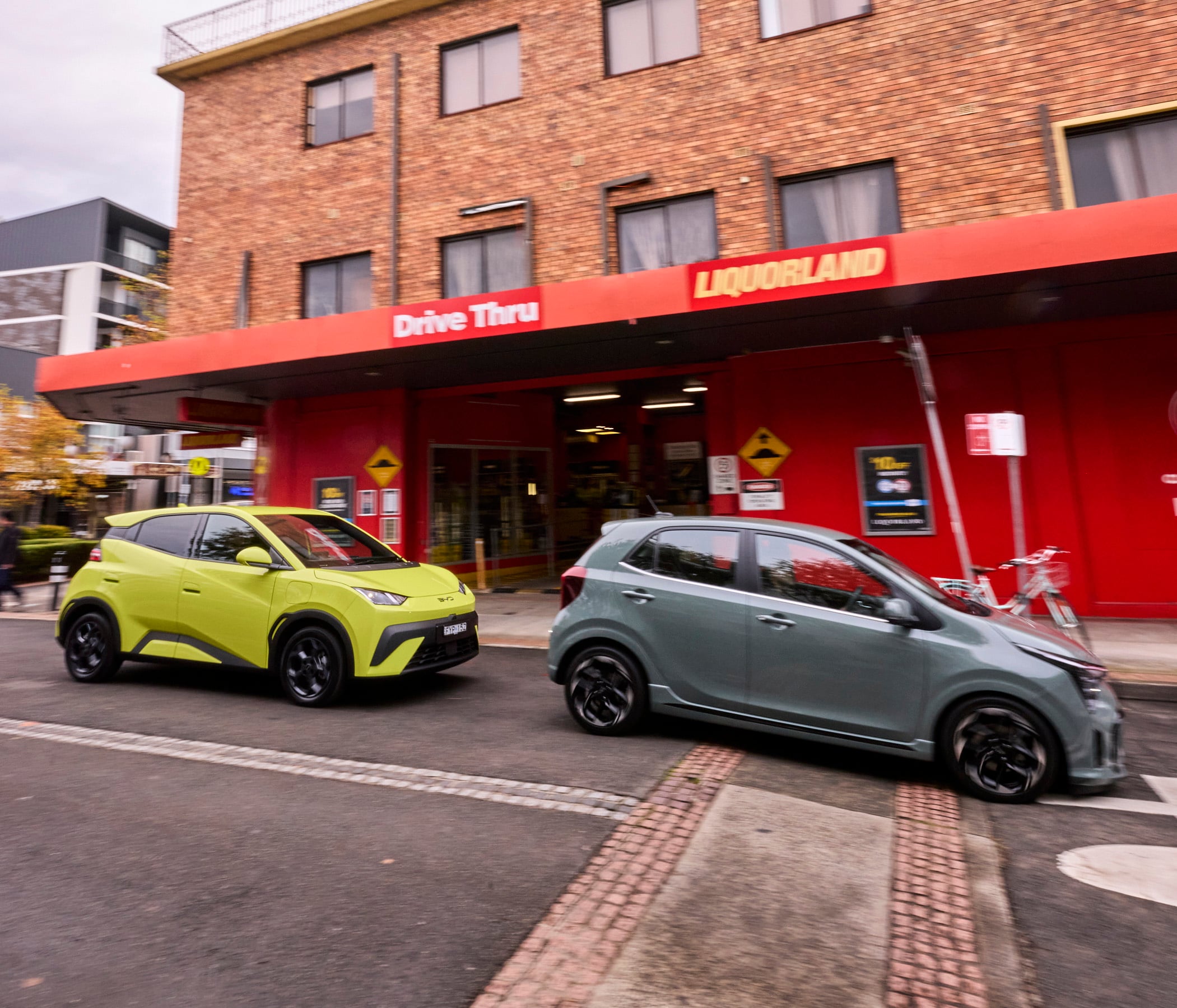 Two cars drive by a red storefront with a 'Drive Thru' sign and 'LIQUORLAND' on a brick building, motion blurred by speed.