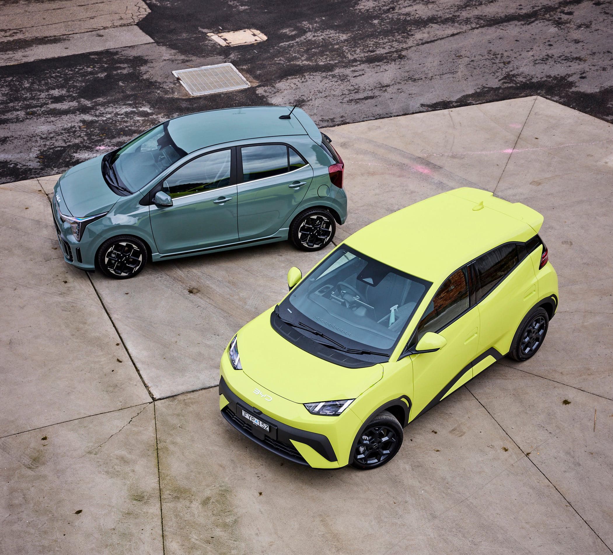 Aerial view of two cars parked diagonally: a teal hatchback in the foreground and a neon yellow car beside it on a gray parking lot.