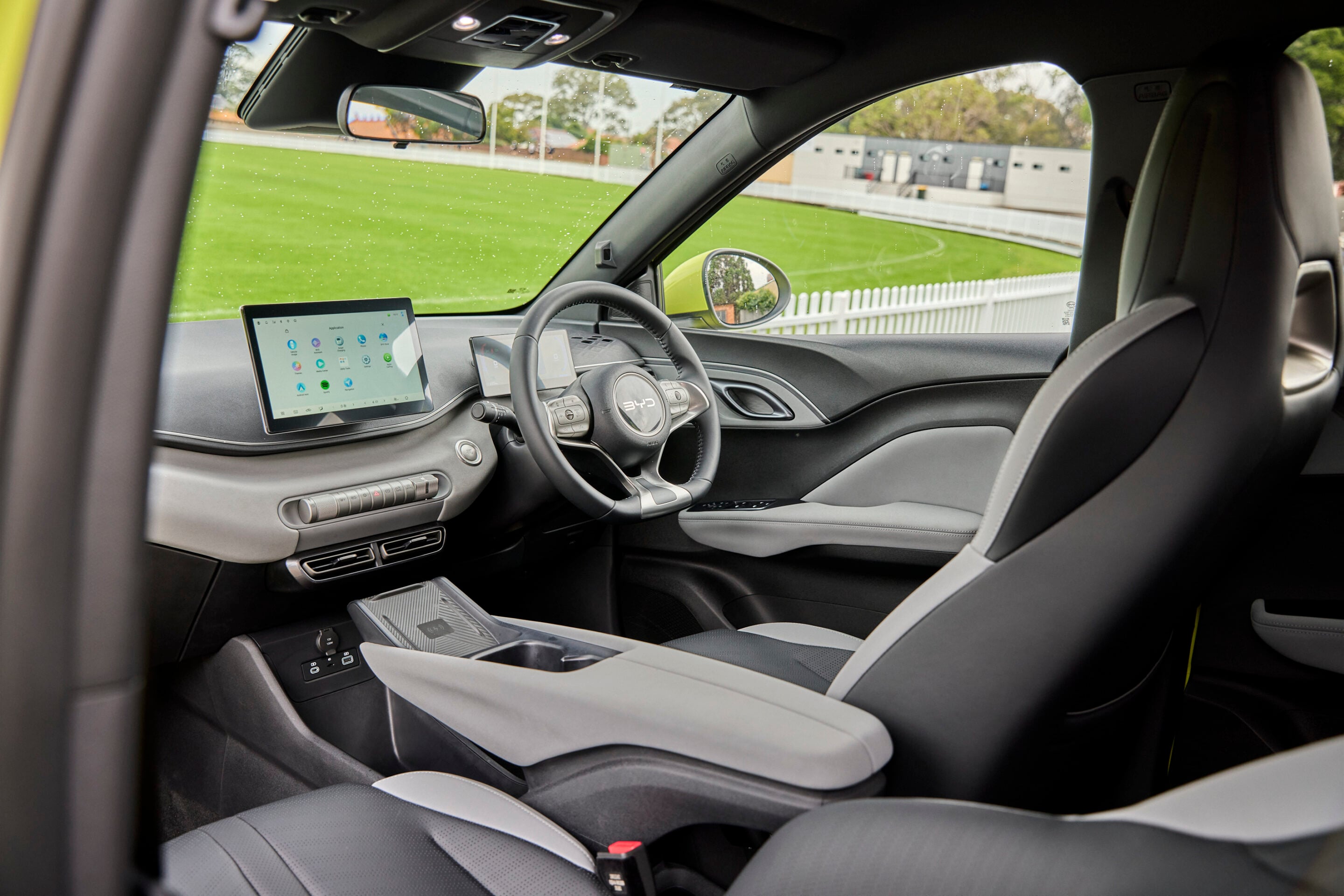 Interior of a modern car showing a large touchscreen dashboard, steering wheel, and gray seats with a green field outside.