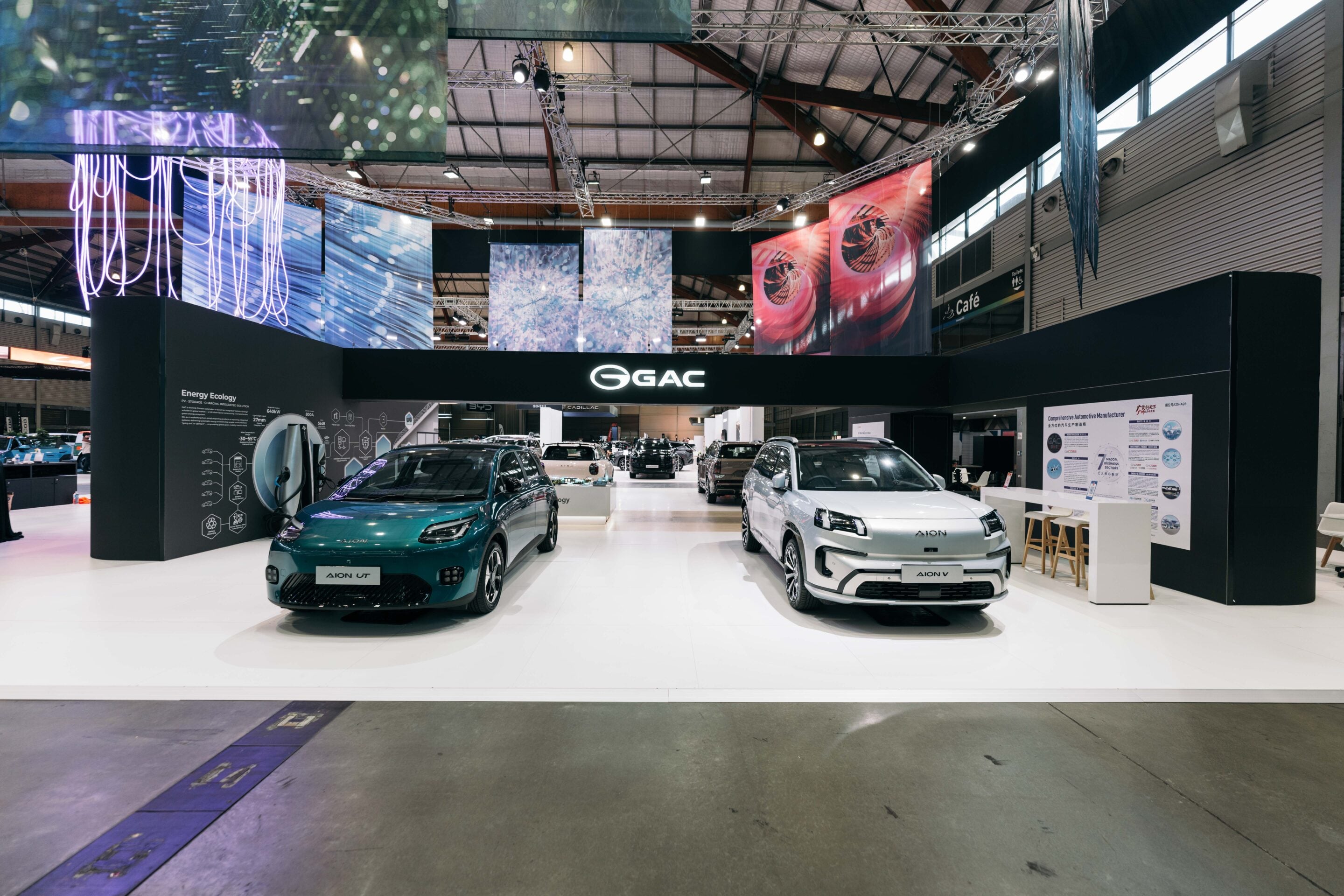 Two electric cars on display at a trade show booth with a black GAC sign and colorful banners overhead.