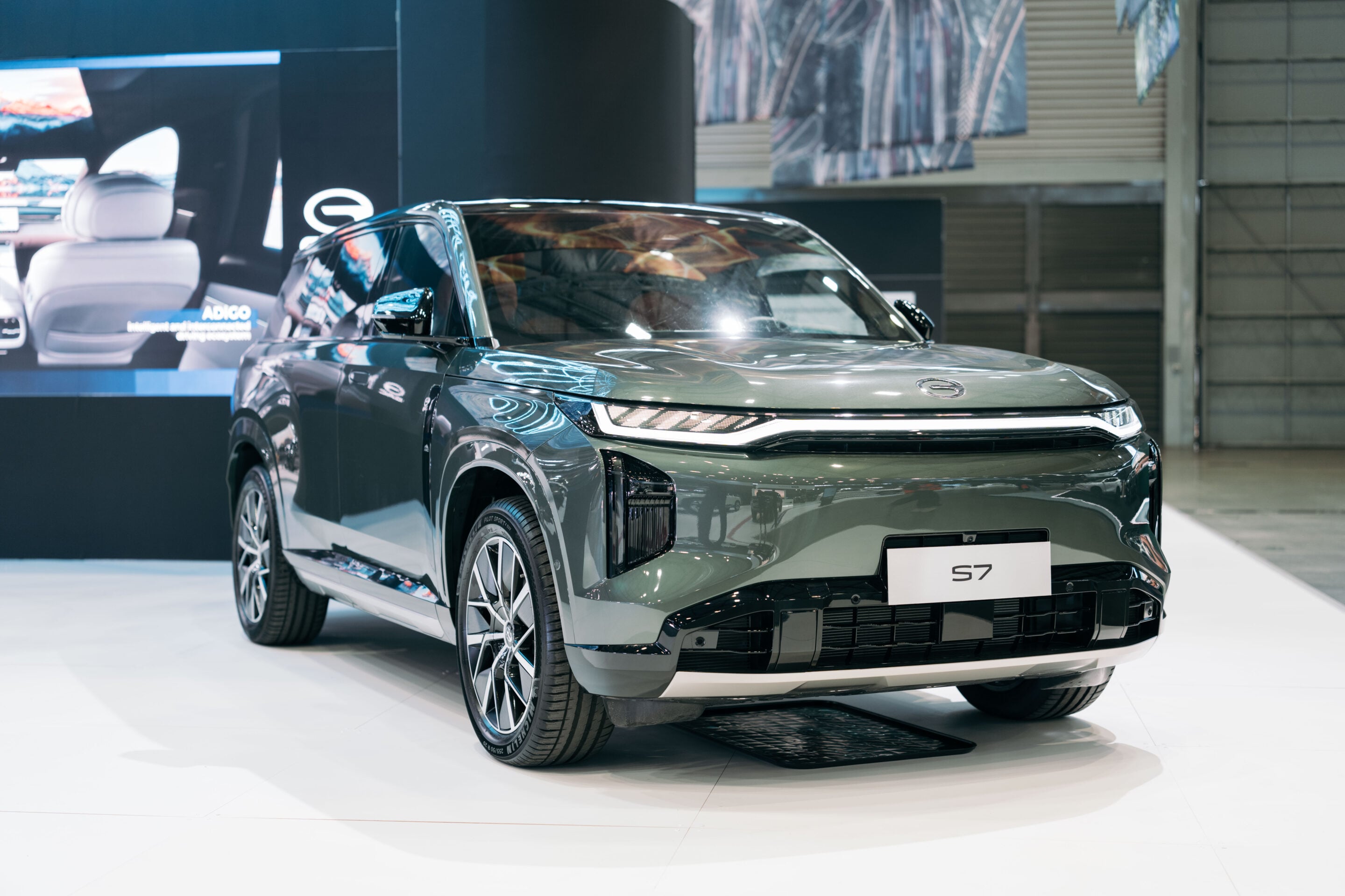 Front three-quarter view of a metallic gray SUV (S7) on display at an auto show, with bright showroom lighting.