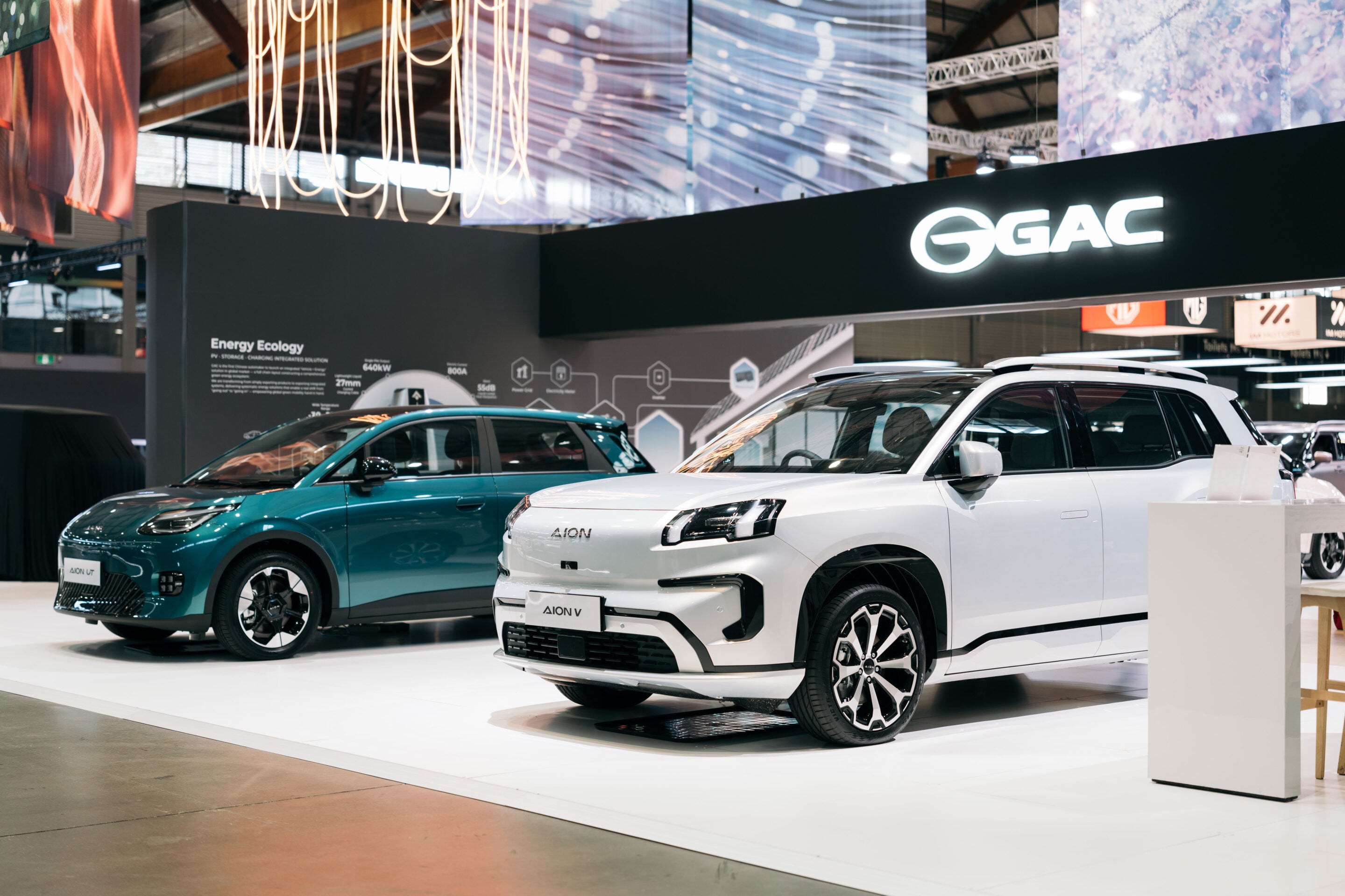 White SUV and teal car on a white exhibition platform under a prominent GAC display at an indoor auto show.