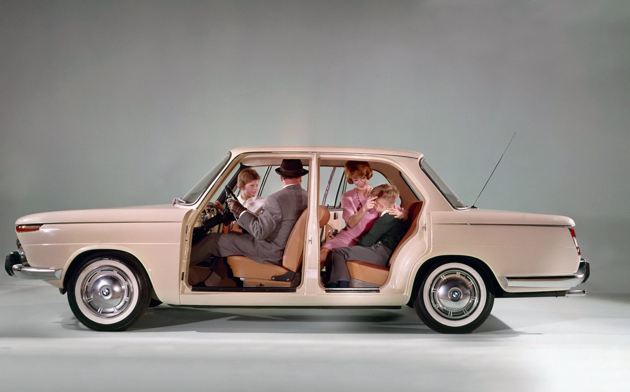 Family of four sits inside a vintage beige car with front doors open, posing as if posing for a photo in a studio setting.