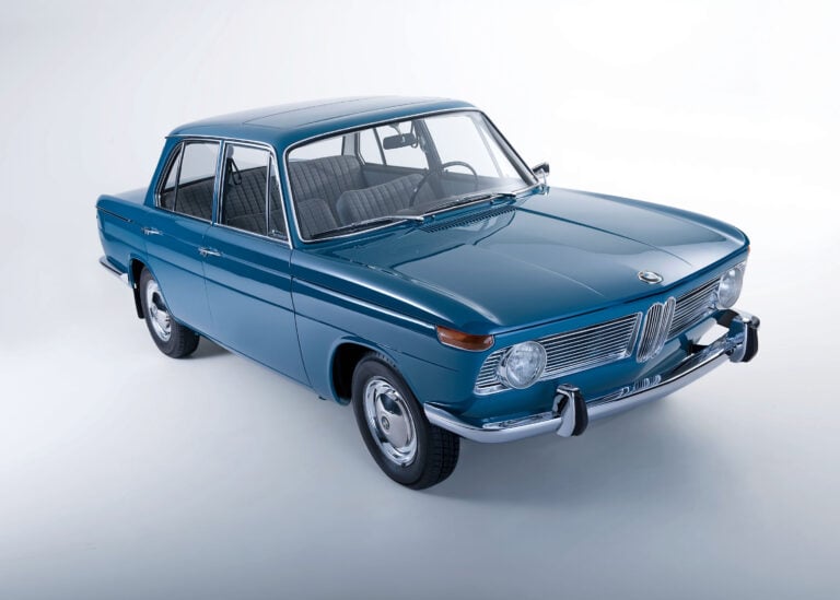 Vintage blue BMW sedan photographed from a front-right angle in a bright studio, showcasing chrome grille and classic rounded headlights.