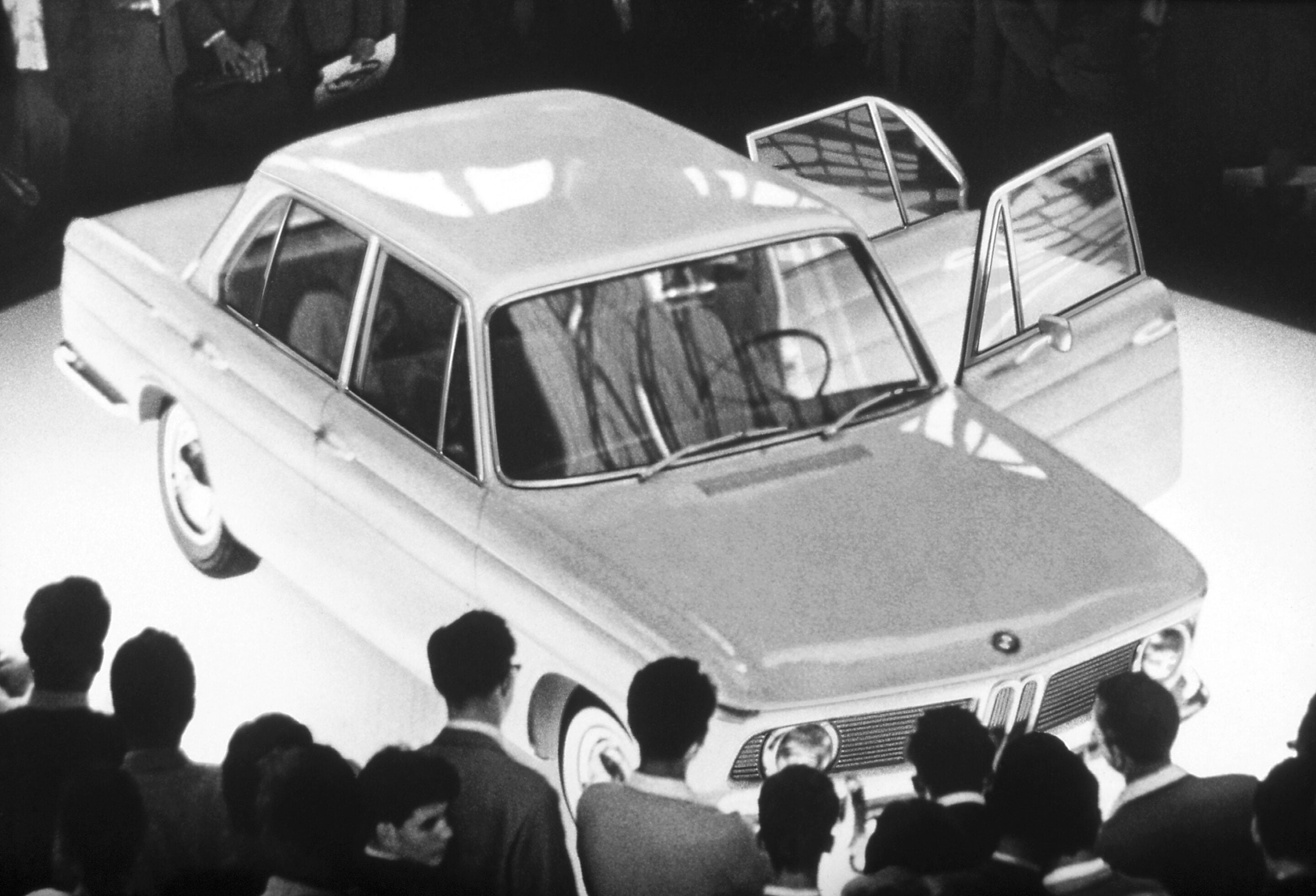 Crowd of people gathered around a vintage car with its doors open at what looks like a public event, black-and-white photo.
