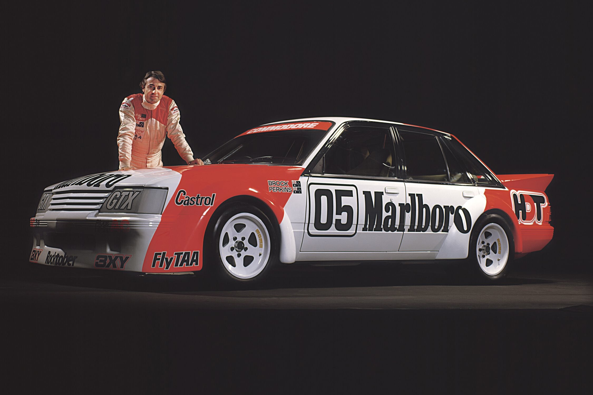 Racer in a white-red suit leans on a red-and-white Marlboro-sponsored race car against a dark background.