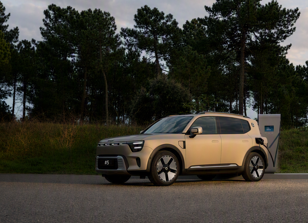 Beige electric SUV plugged into a roadside charging station with pine trees in the background.