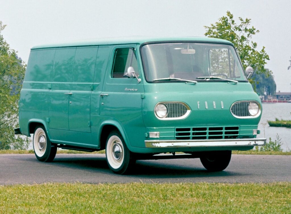 Front of a vintage mint-green Ford van with round headlights parked in a lot, trees in the background.