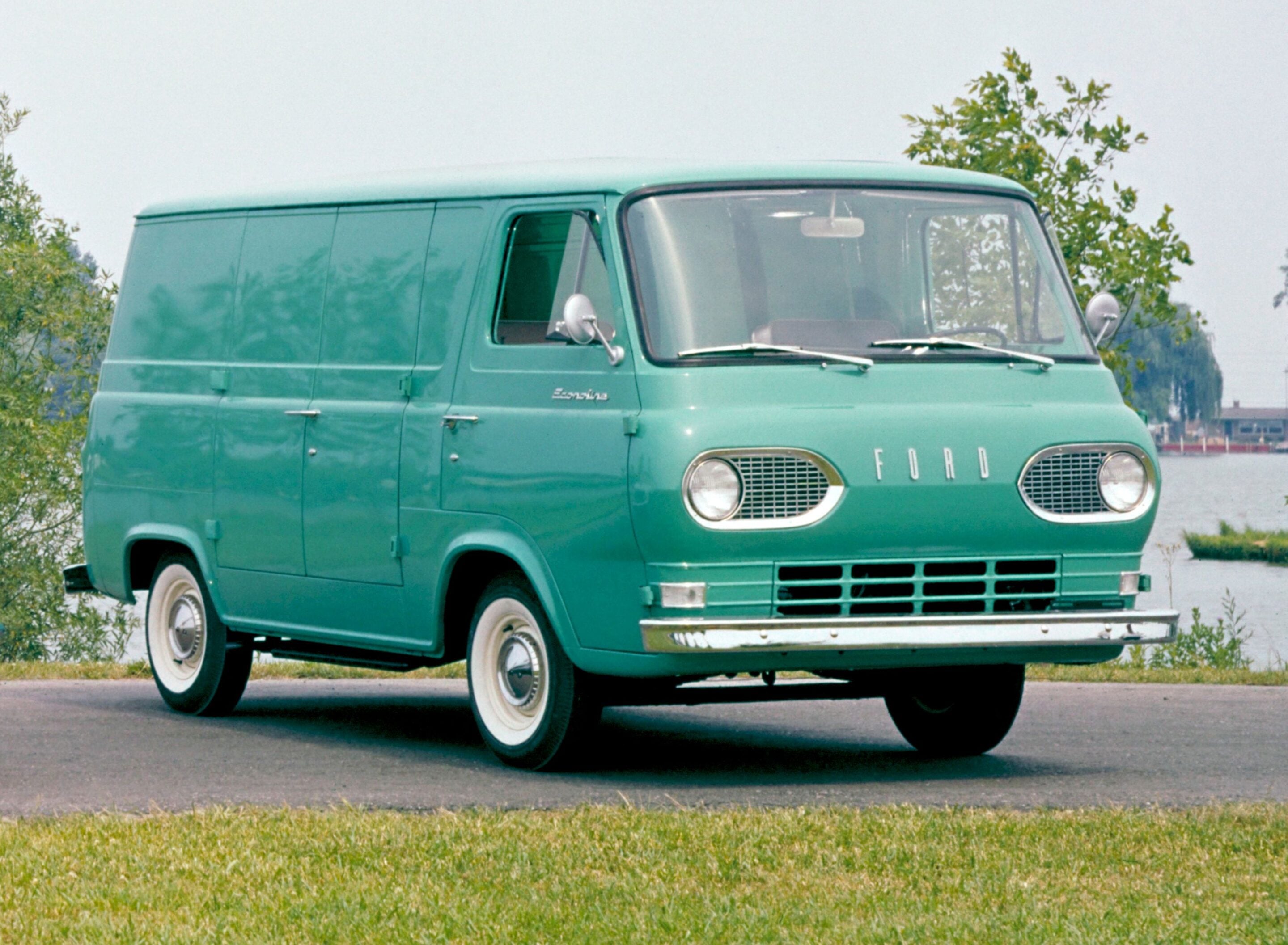 Front of a vintage mint-green Ford van with round headlights parked in a lot, trees in the background.