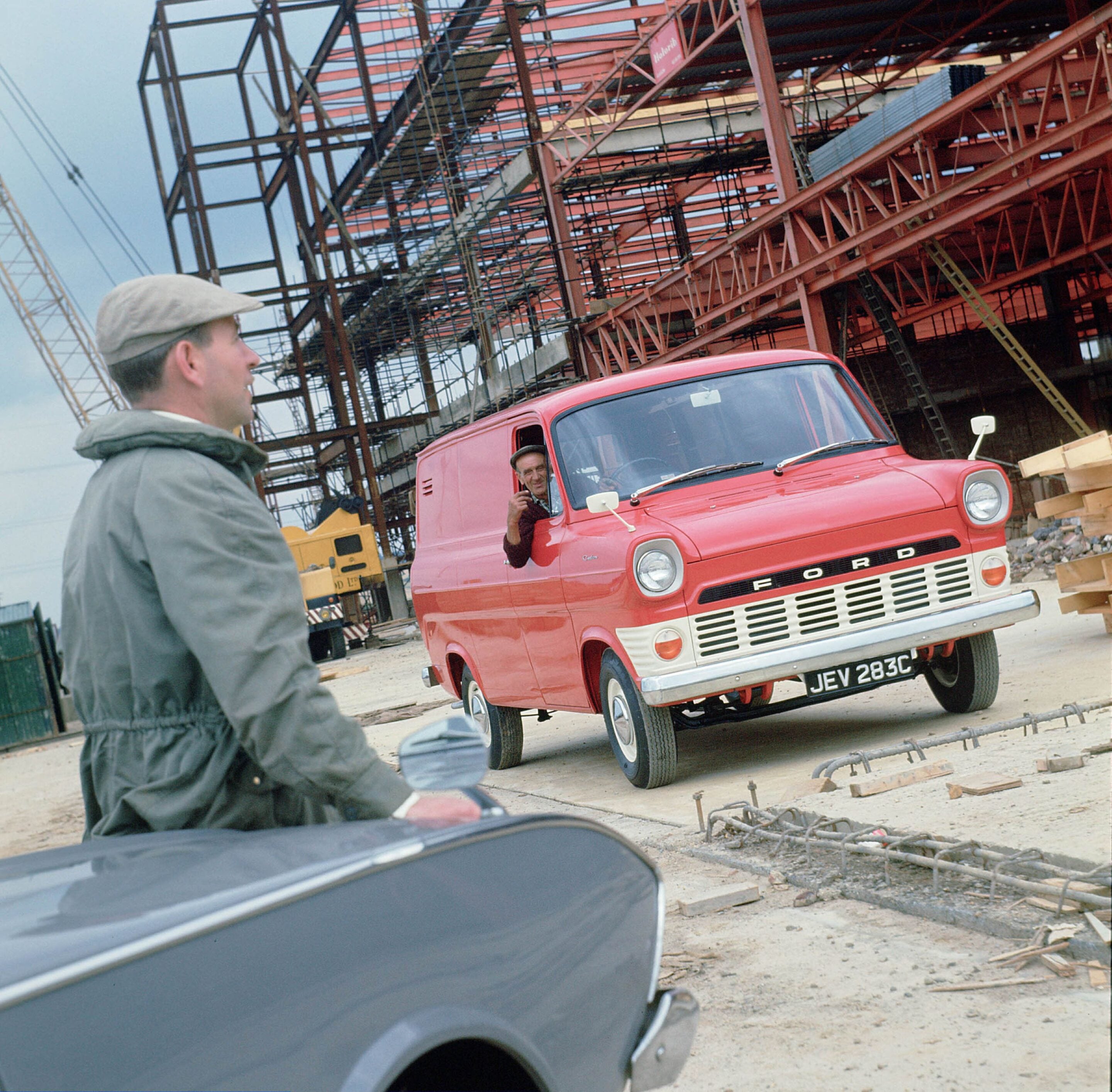 Construction site with a red vintage Ford truck and workers, one man in a gray jacket in the foreground and another in the truck cab nearby.