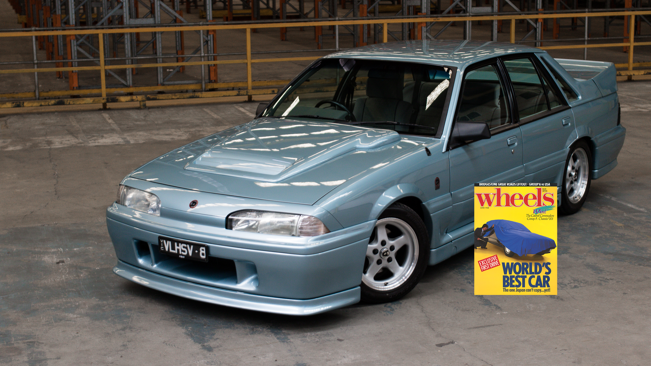 Light blue sedan parked in an industrial garage, with a colorful 'Wheels' magazine display beside its rear door.