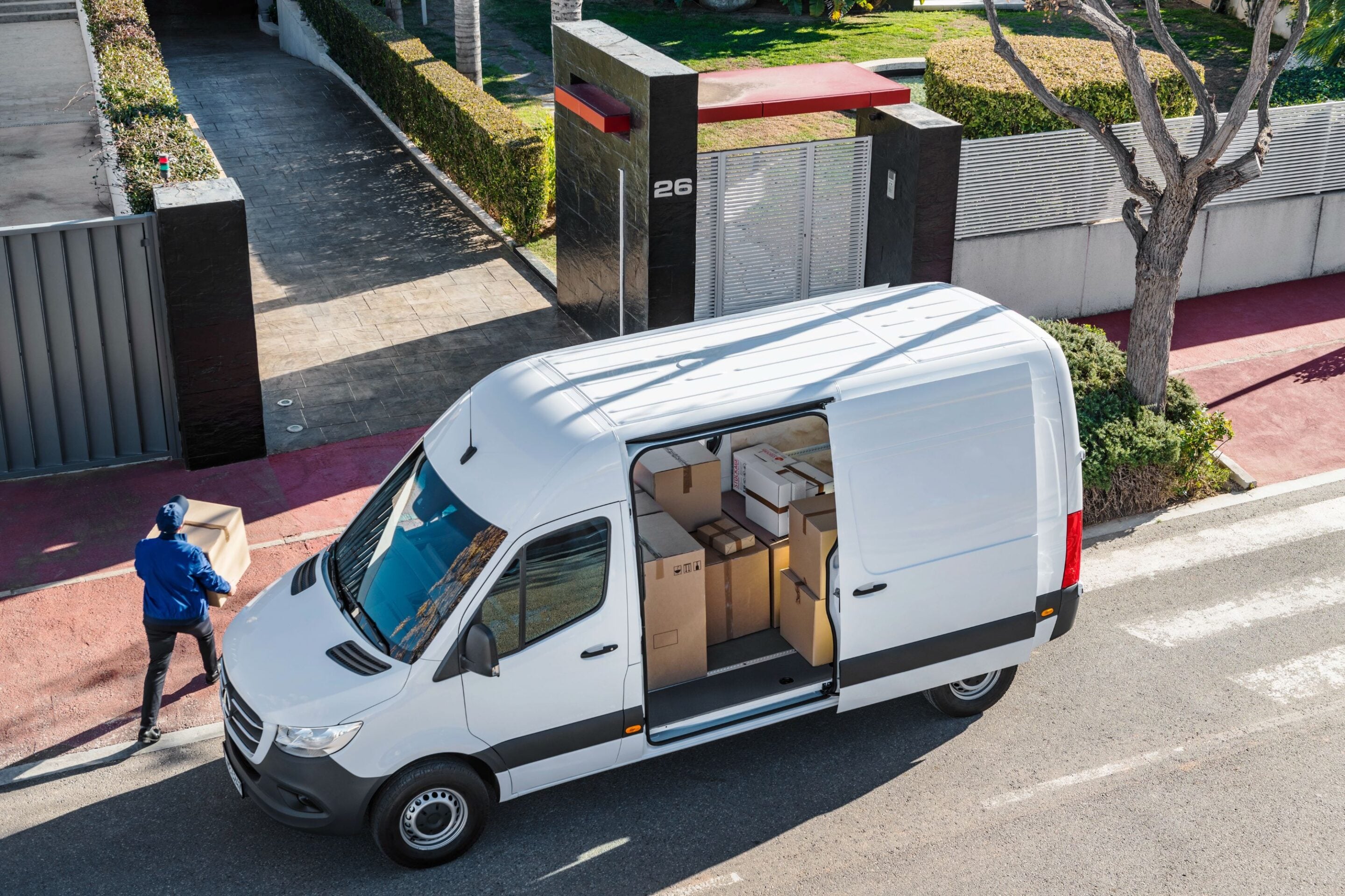 Overhead view of a white delivery van with its cargo door open, stacked with cardboard boxes, as a worker unloads nearby.