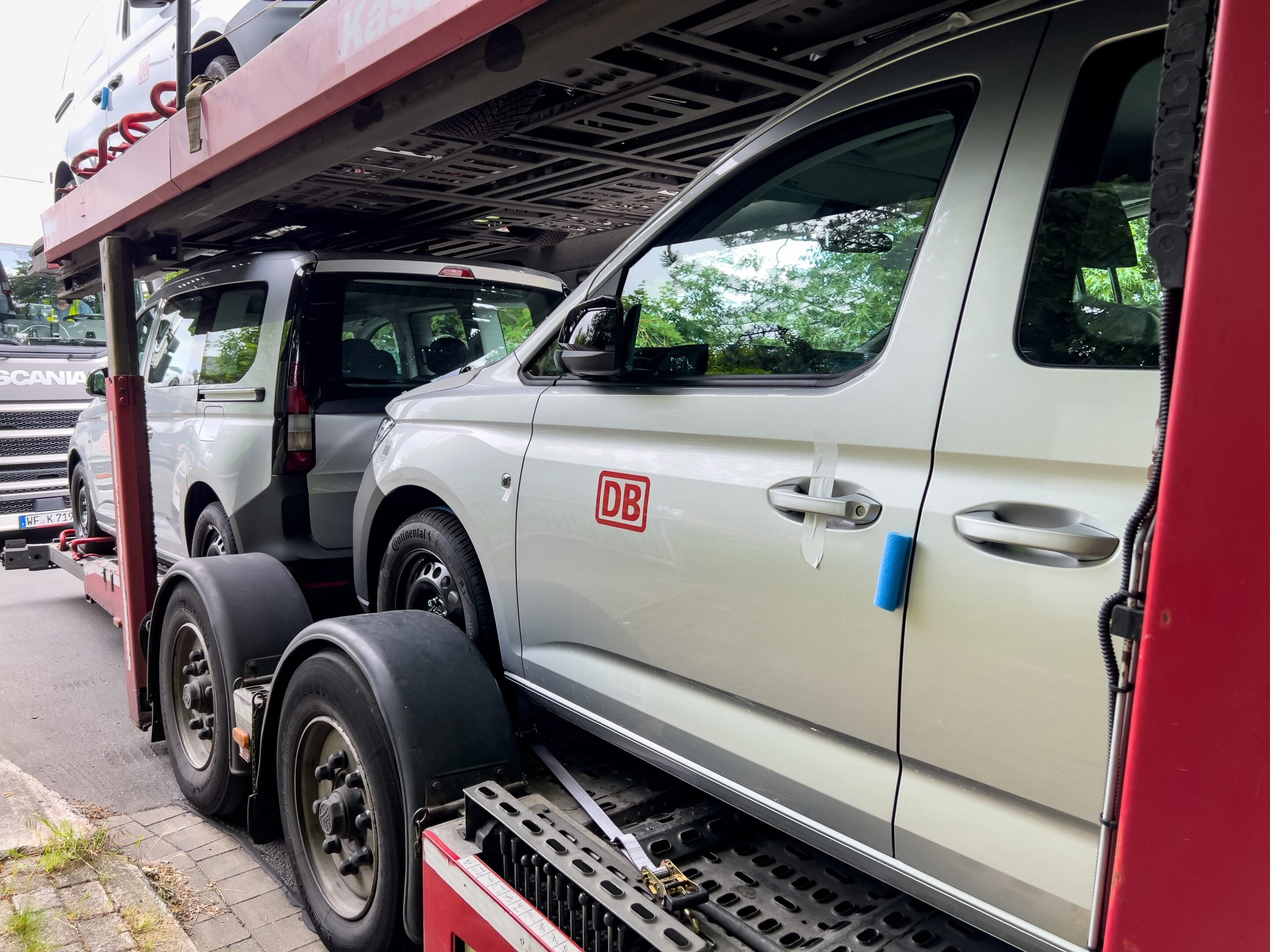 White van loaded on a car transport trailer with another vehicle above, red trailer arms, and a DB logo visible on the side