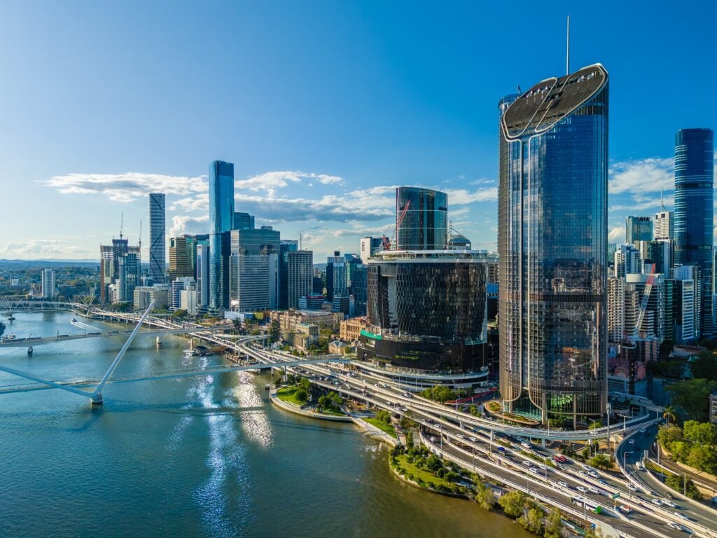 Aerial view of a modern downtown waterfront with glass skyscrapers, a curving bridge, and multiple highways along the river under a clear blue sky