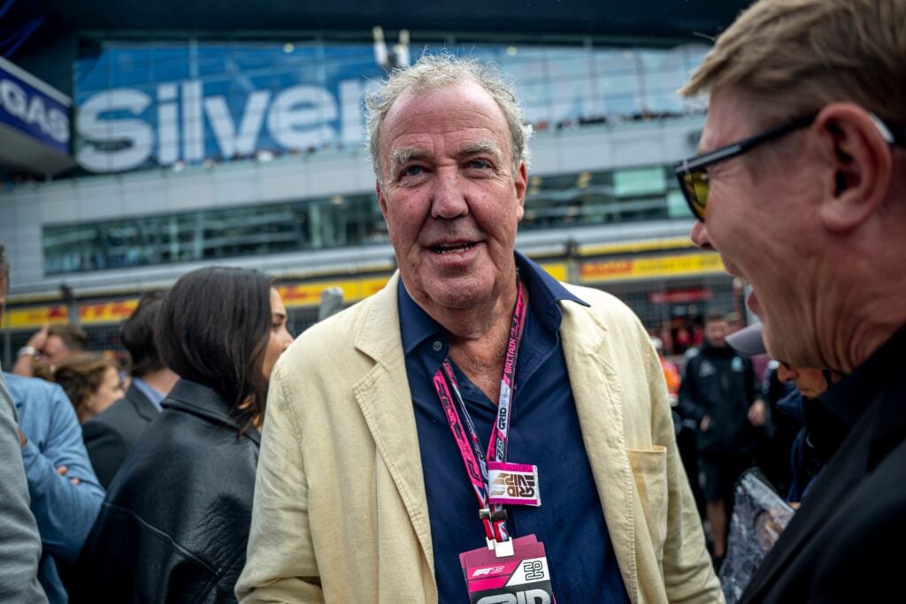 Man in a beige blazer with a pink lanyard at a crowded event, backdrop shows a modern building and rail barrier.