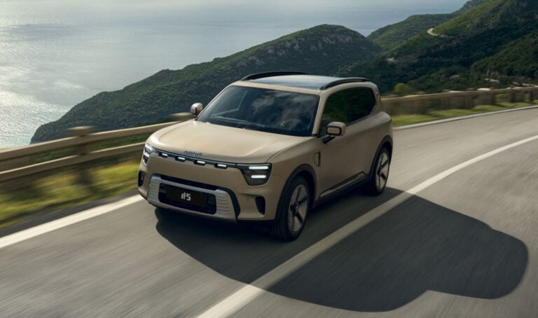 Beige compact SUV driving along a coastal mountain road with the sea to the left and green hills in the background.