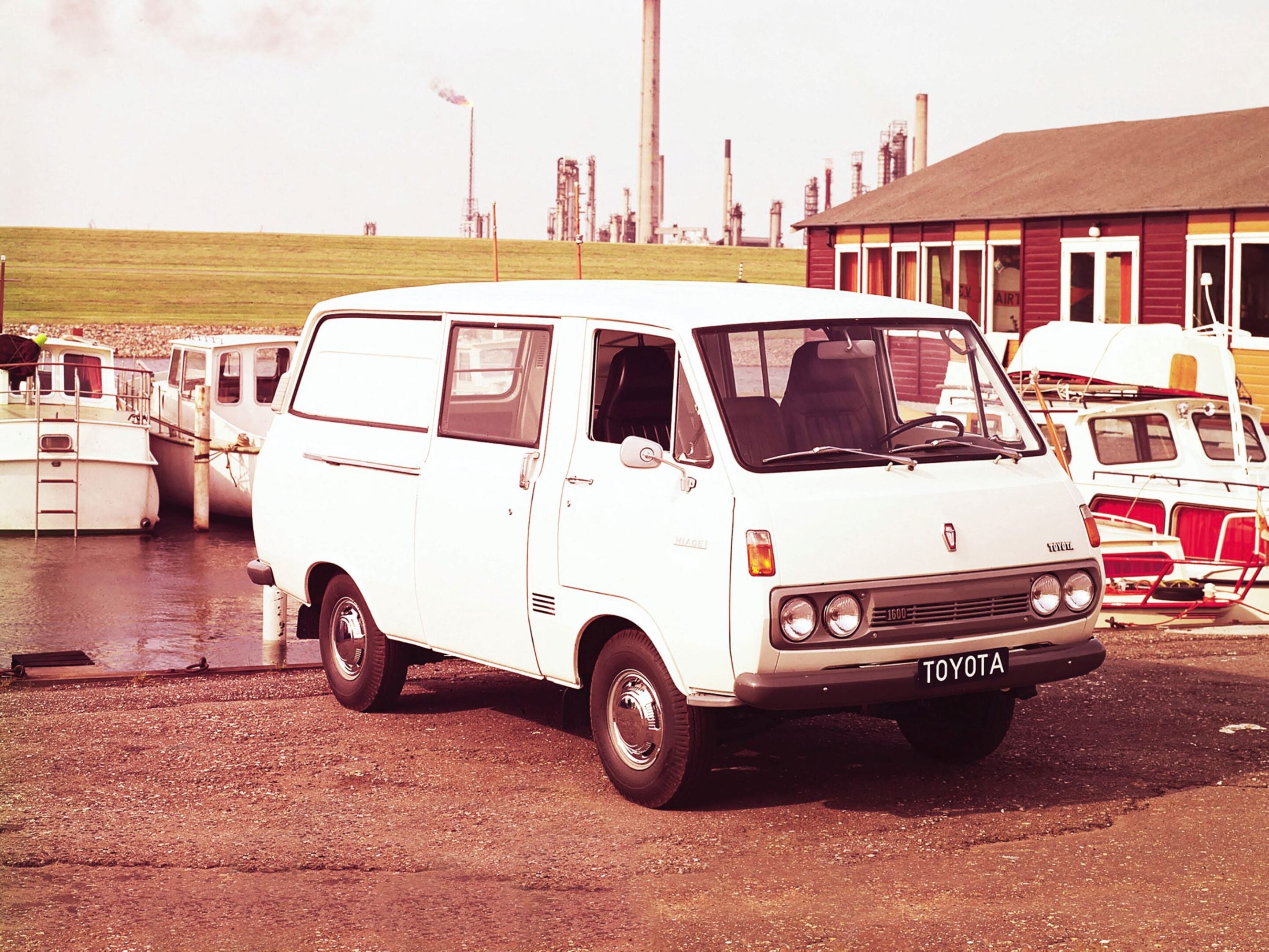 White Toyota van in a marina foreground with moored boats and a red wooden building, and industrial towers visible in the distance.