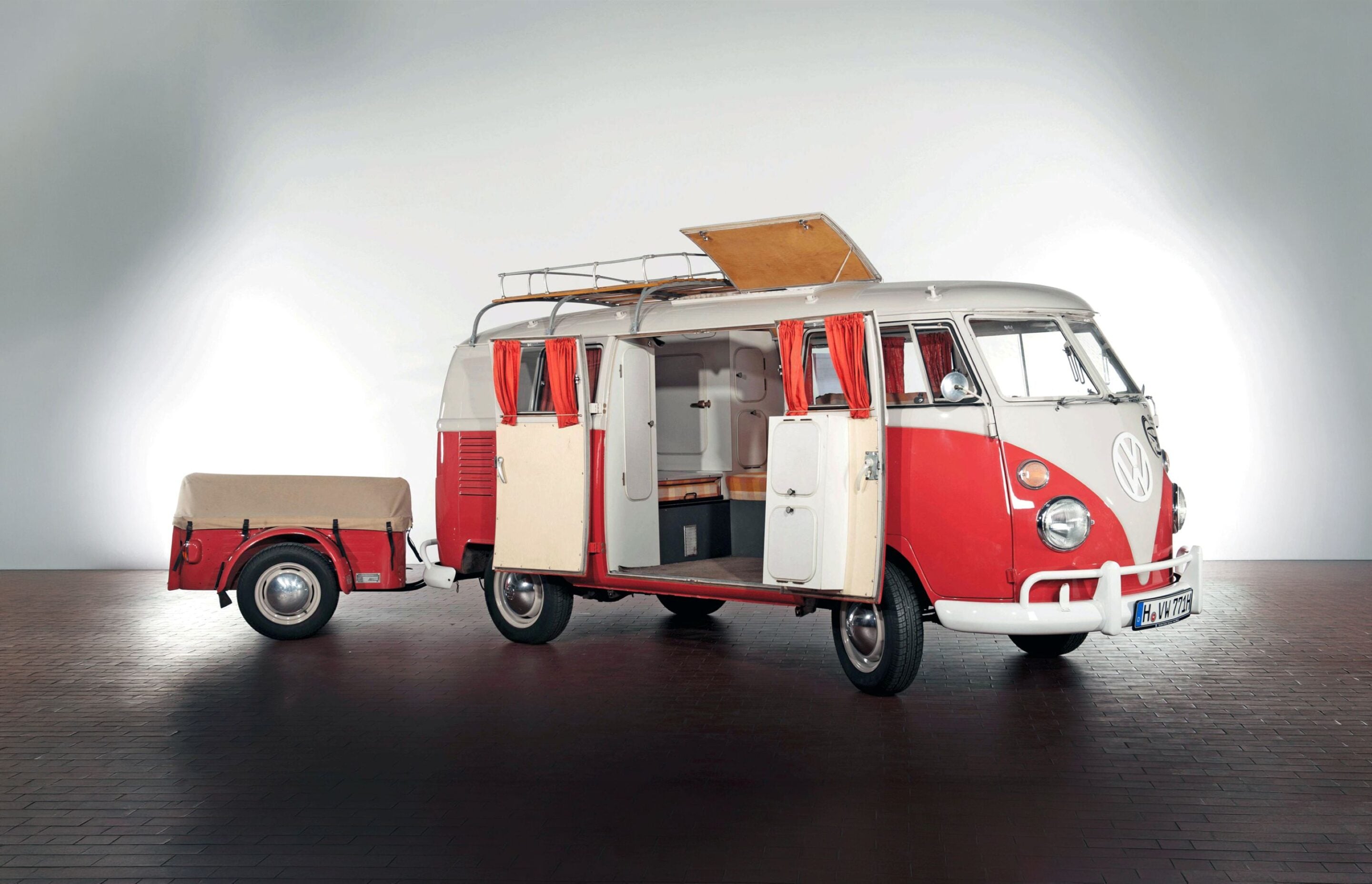 Vintage red-and-white VW camper van with open doors and a raised pop-up roof, hitched to a small matching trailer in a studio setting.
