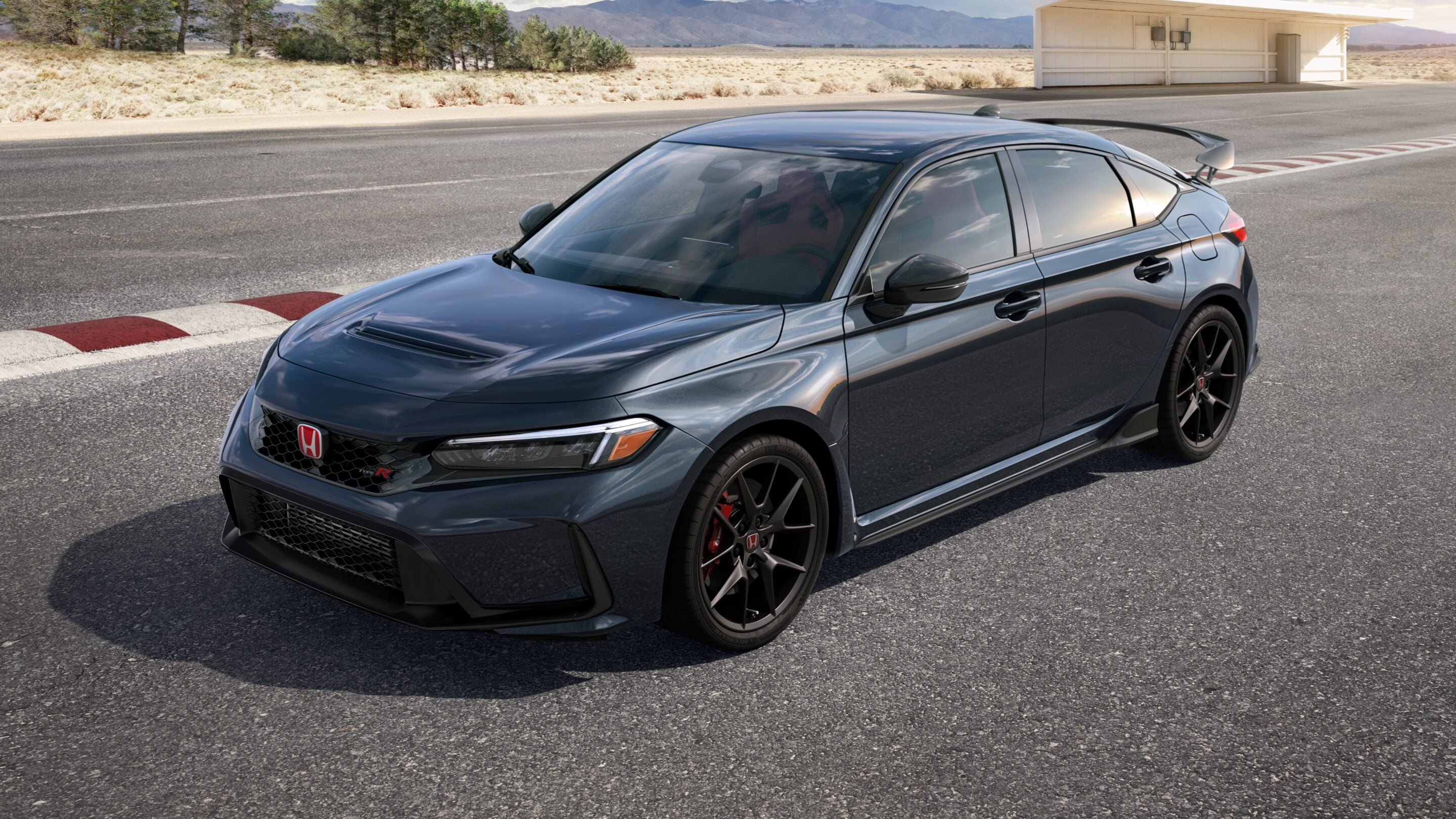 Grey Honda Civic Type R sports sedan on a desert highway with a hangar and distant mountains in the background, sunlit.