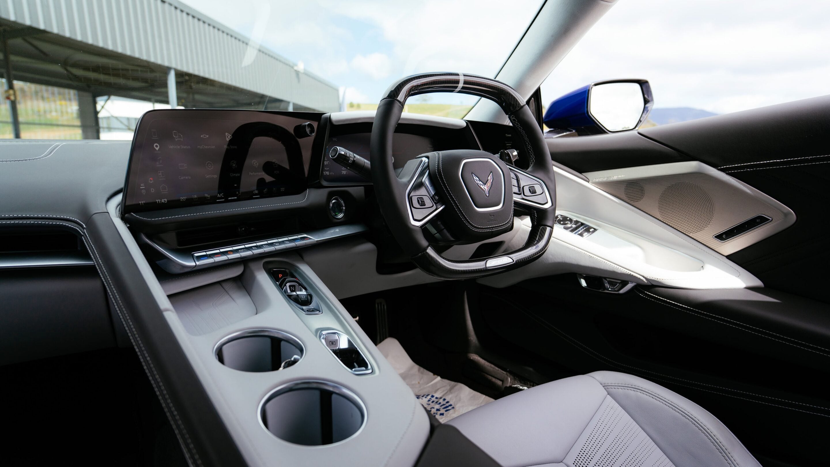 Steering wheel with a Corvette emblem in a premium car interior, featuring a large curved touchscreen infotainment panel and stitched leather.