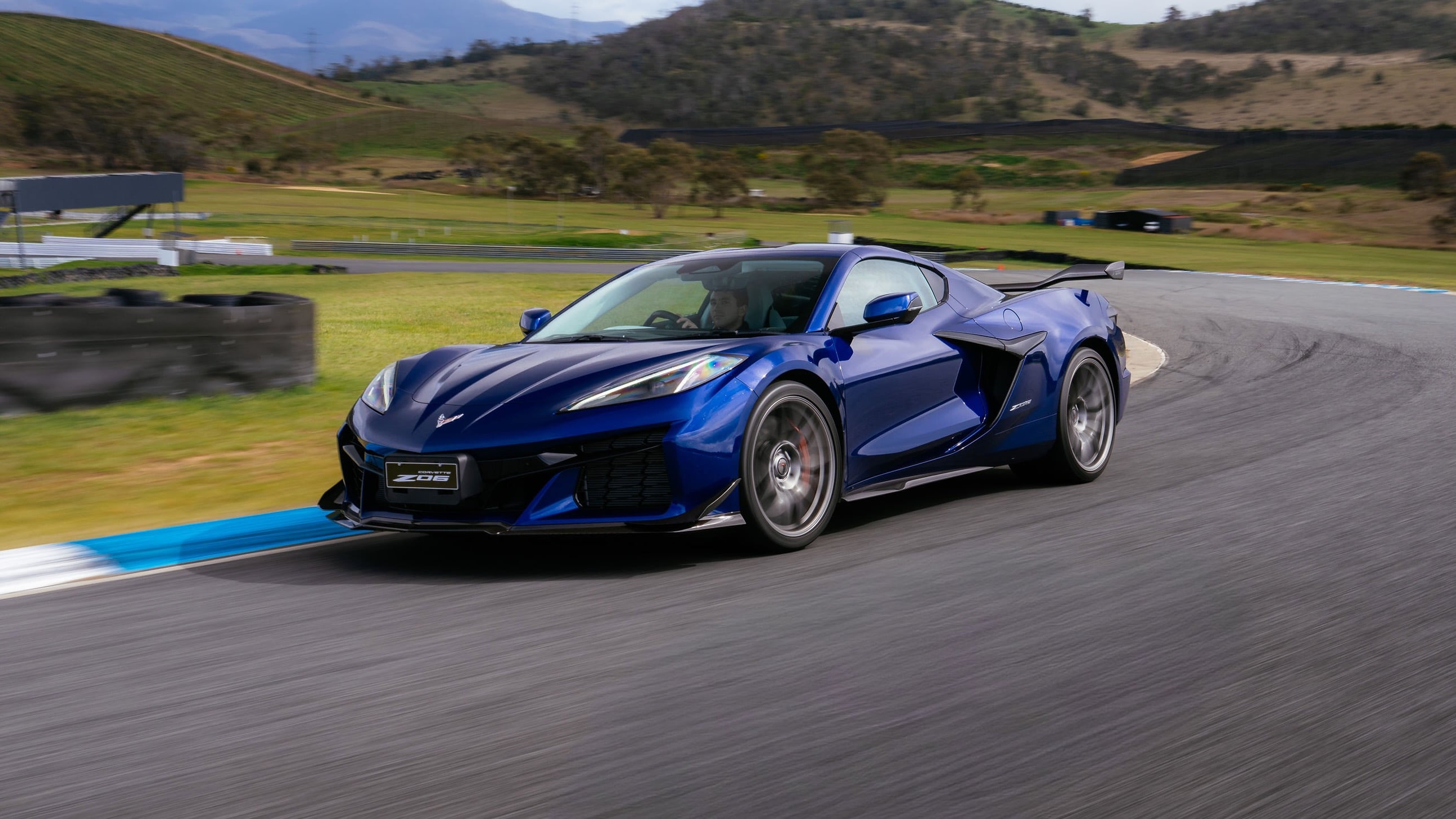 Blue Corvette C8 speeding through a racetrack hairpin, motion blur on tires, green hills in background