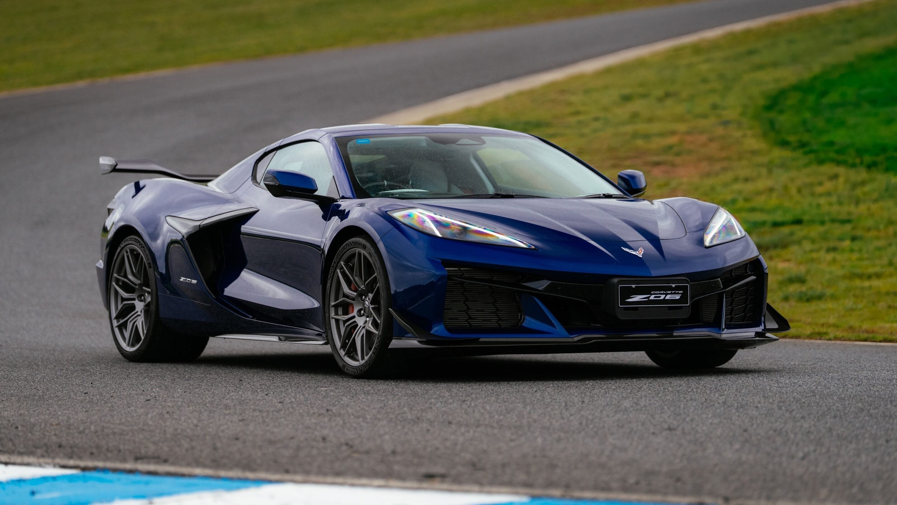 Blue Chevrolet Corvette Z06 on a race track, photographed from the front-left angle with curving pavement and grass in the background.