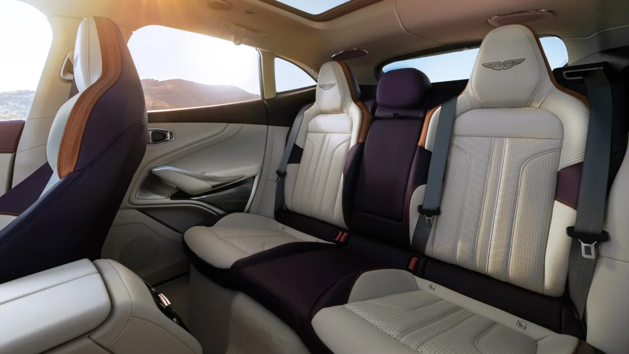 Luxurious rear car seating with light leather upholstery, two outer seats and a center seat, Aston Martin logos on headrests, sunlit window nearby, seat belts fastened.