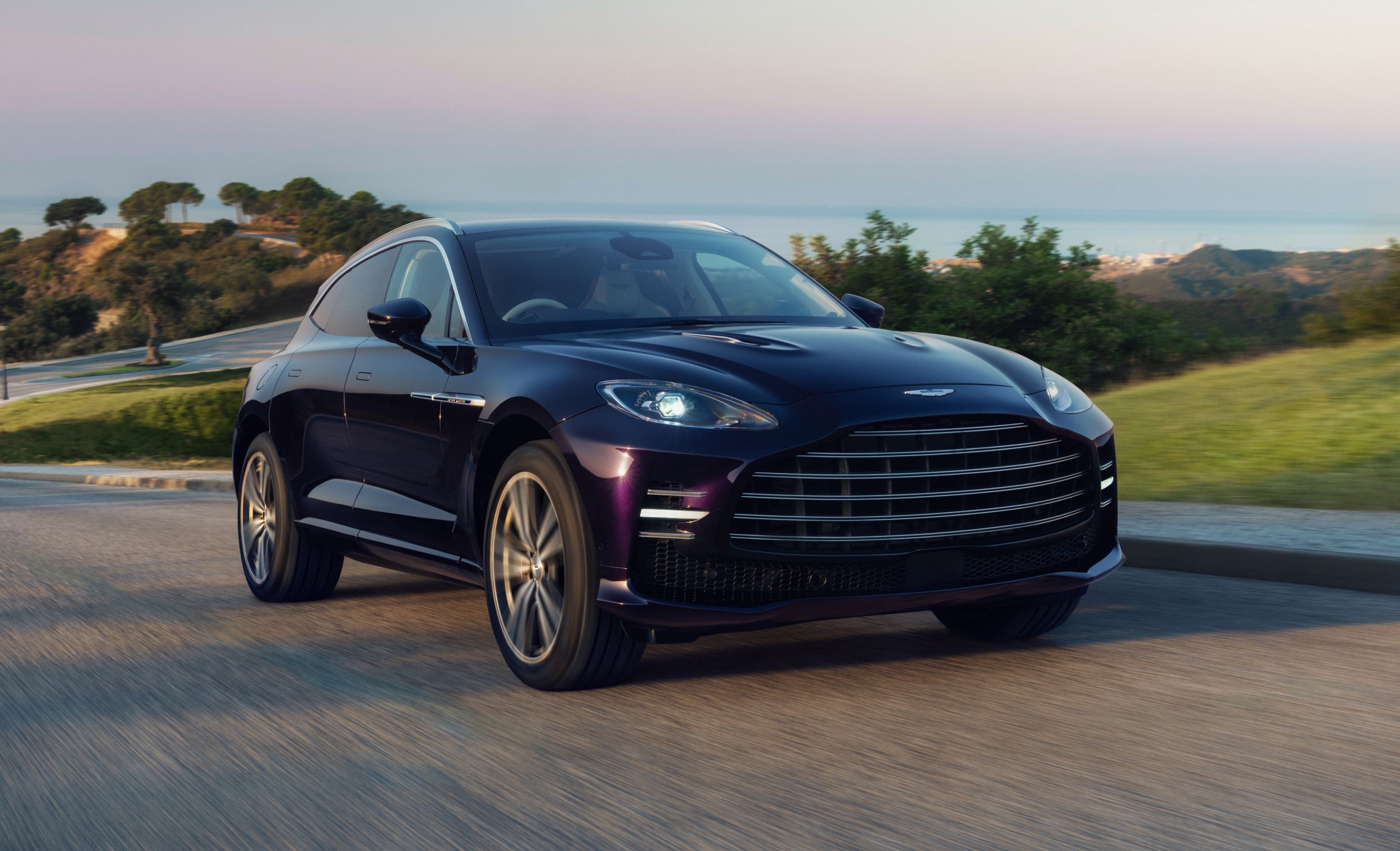 Dark blue luxury sedan speeding along a coastal highway with hills and the sea in the distance.