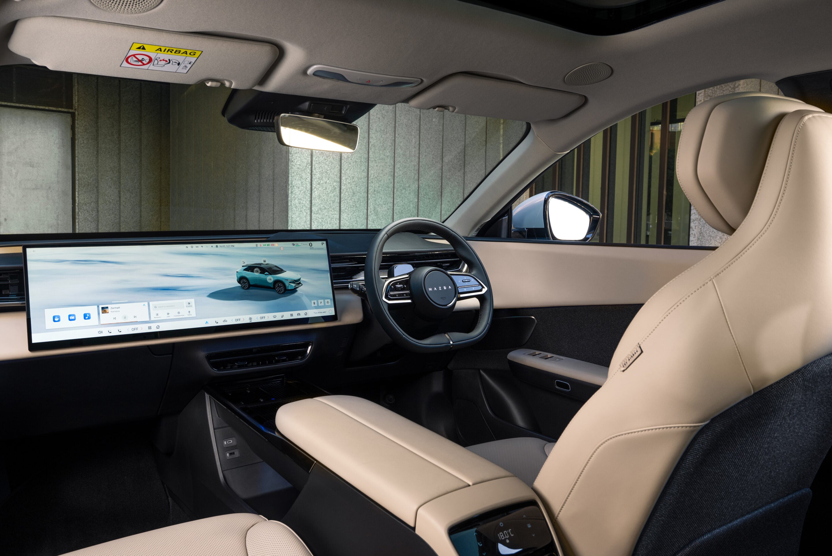 Inside a luxury car: beige leather seats and a wide horizontal touchscreen display across the dashboard with a sporty steering wheel nearby.