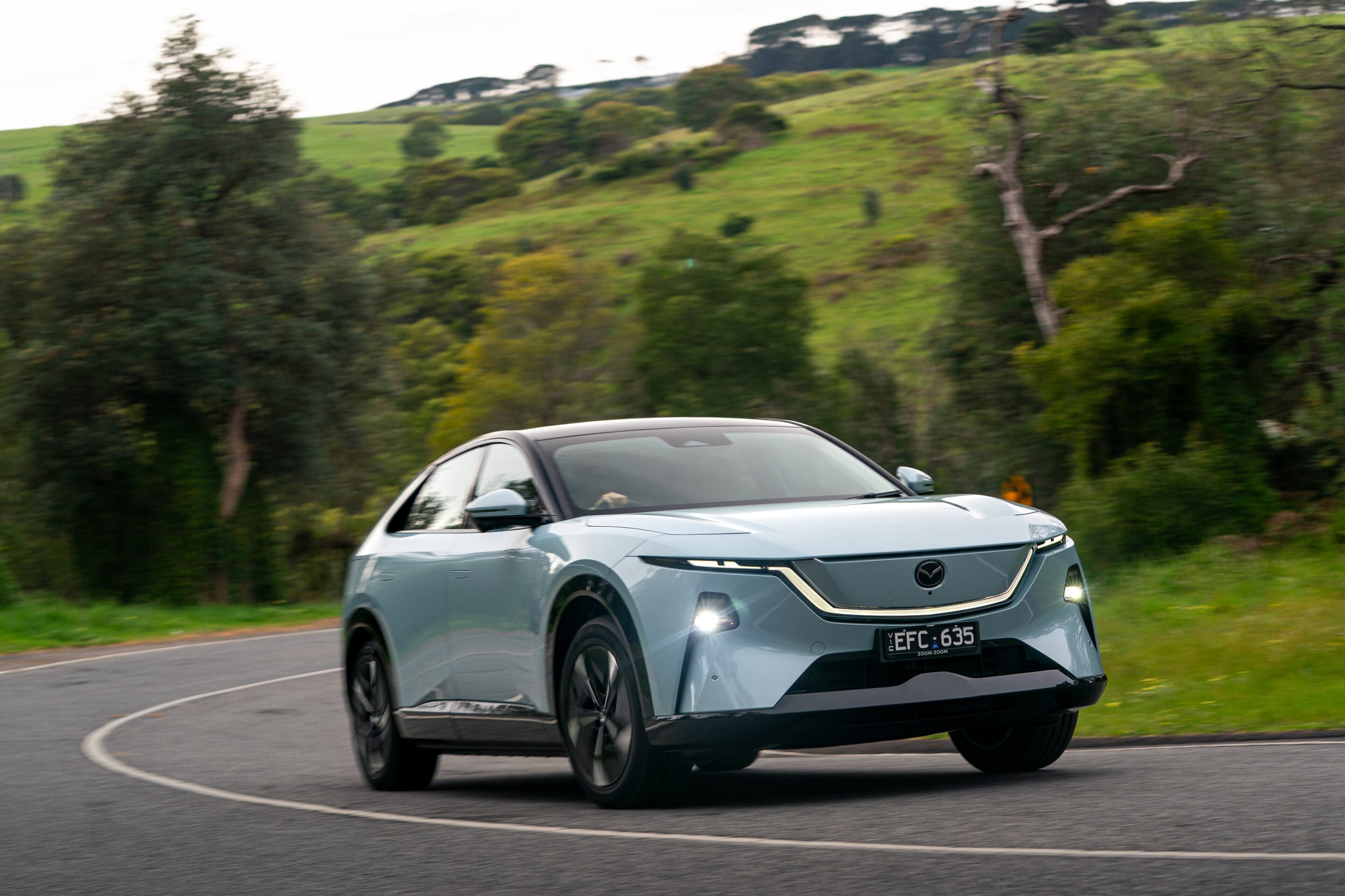 Silver car driving on a curved rural road with green hills and trees in the background, motion-blurred scenery.