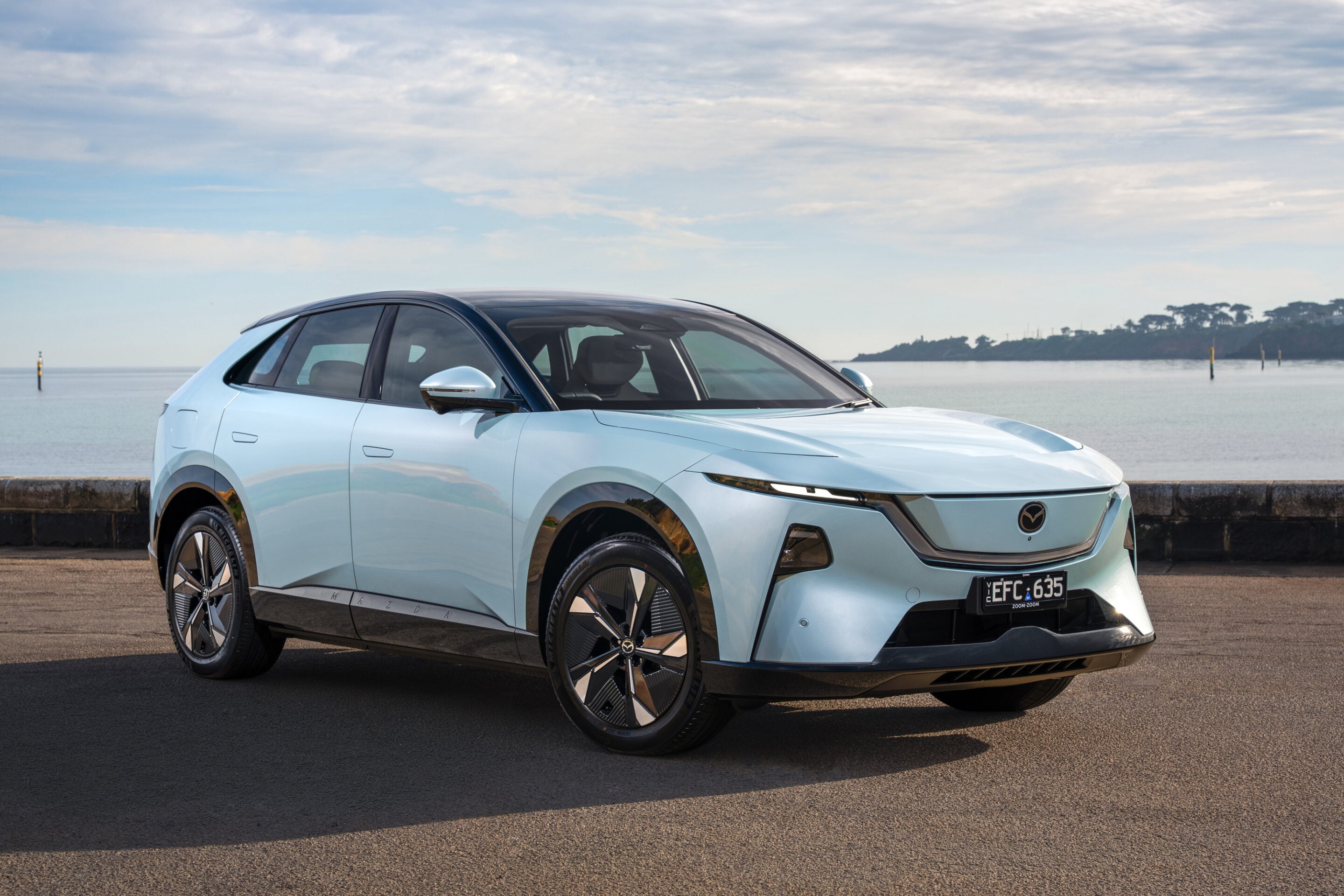 Light blue electric SUV parked by a calm sea with a distant shoreline and cloudy sky in the background.