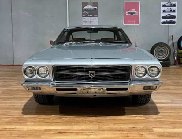 Front view of a light blue vintage car with a chrome bumper, parked indoors on a wooden floor.