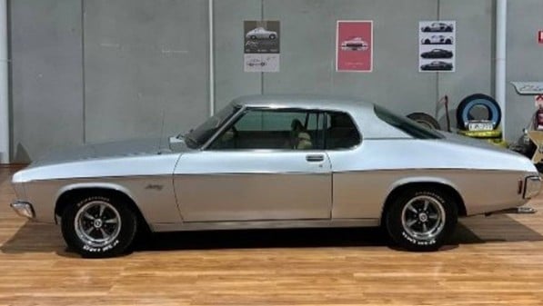 Silver classic two-door coupe on a wooden showroom floor, with posters on the wall behind it.