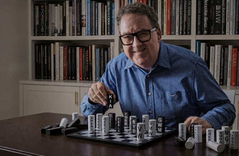 Man wearing glasses and a blue checkered shirt sits at a table, playing Scrabble with letter tiles; bookshelves line the background behind him.
