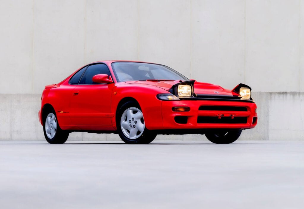 Red Toyota Supra sports coupe with pop-up headlights raised, parked against a pale wall.