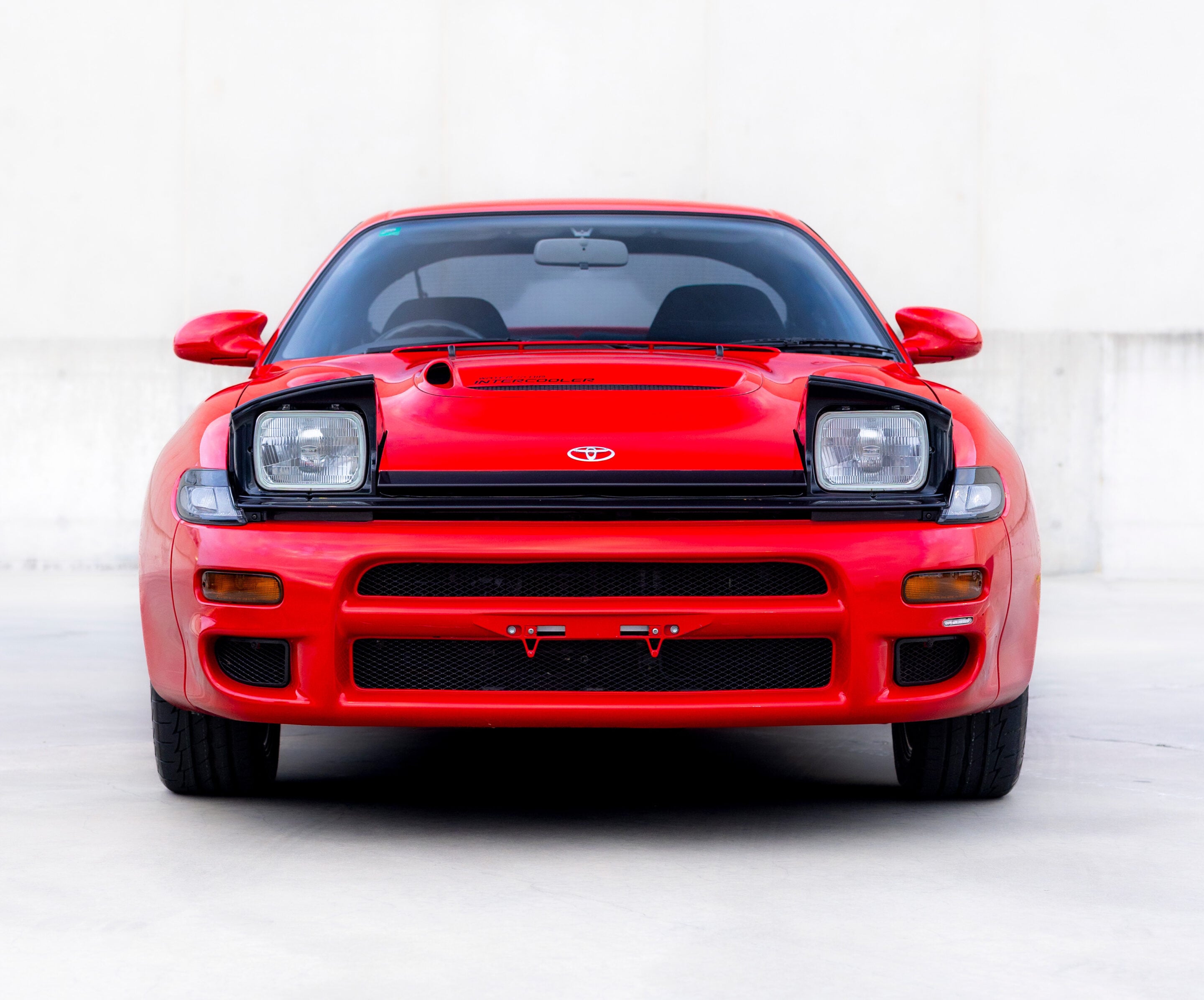 Front view of a red Toyota sports car facing the camera, showing the headlights and grille against a light background.