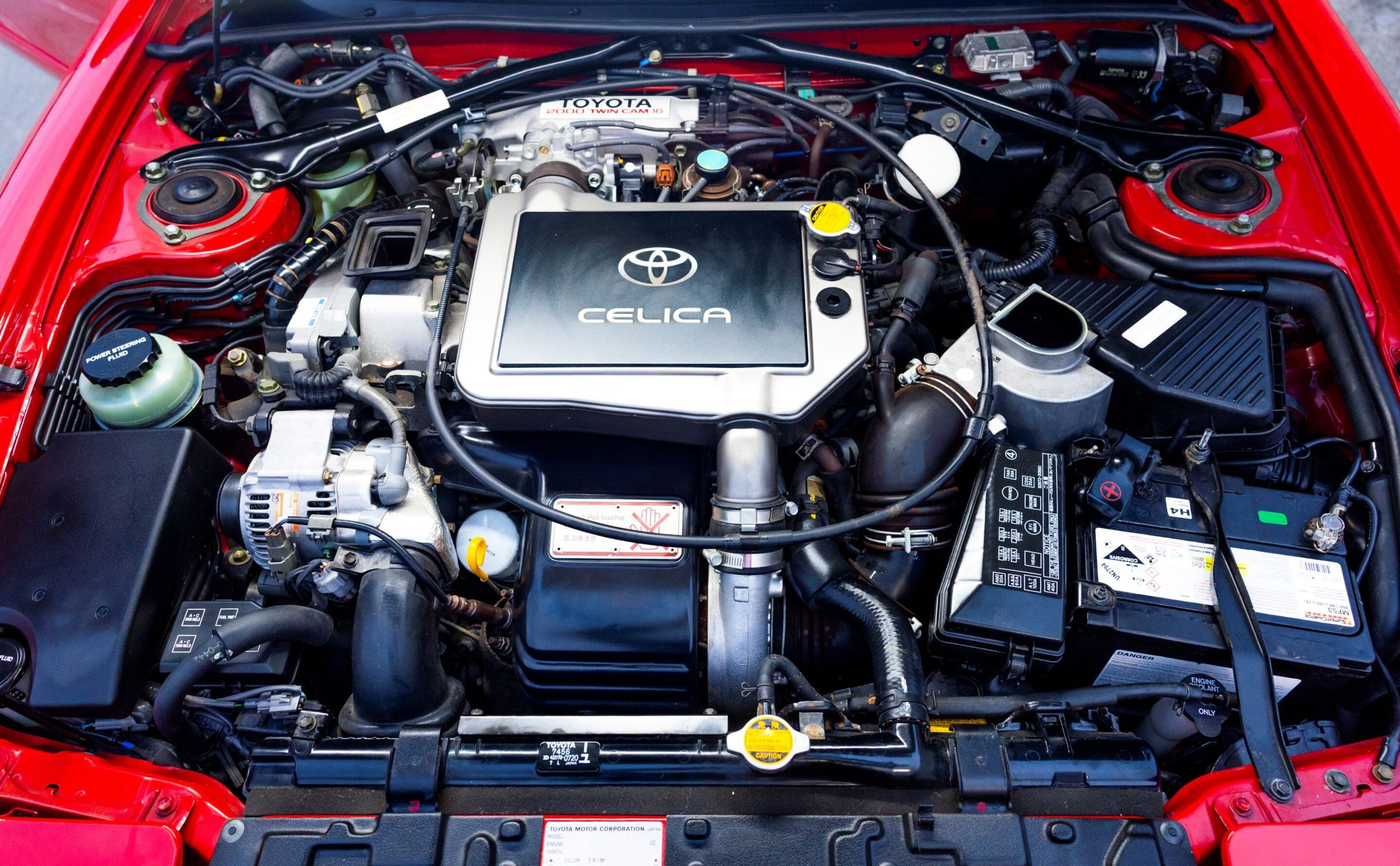 Open engine bay of a red Toyota Celica, showing the silver engine cover with the Toyota logo and 'CELICA' text central to the view.