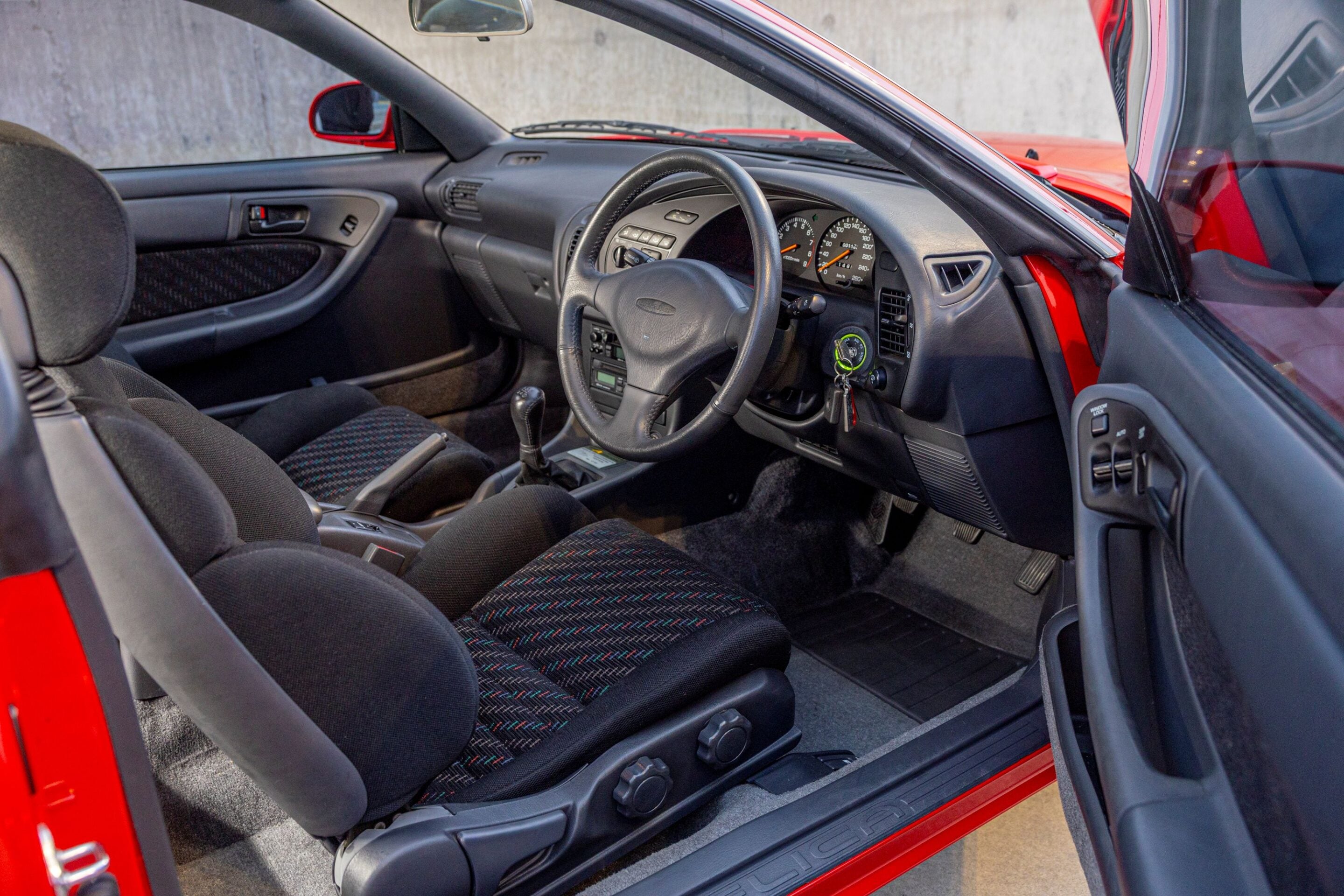Interior of a red car: front seats, steering wheel, and dashboard visible from the passenger side with the driver's door open