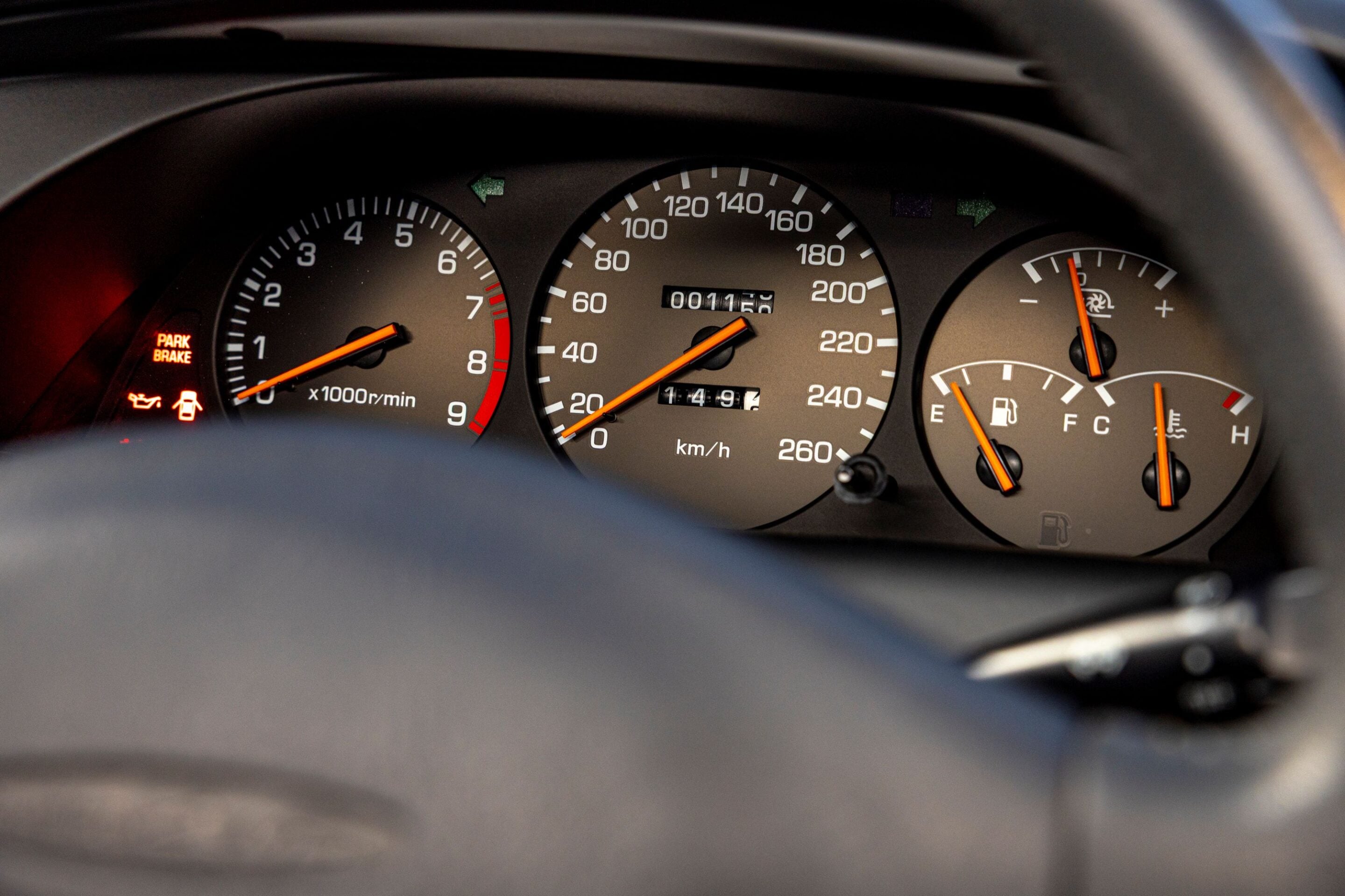 Car dashboard: speedometer up to 260 km/h with orange needle, tachometer on left, fuel and temp gauges on right, illuminated warning icons nearby (Park Brake).