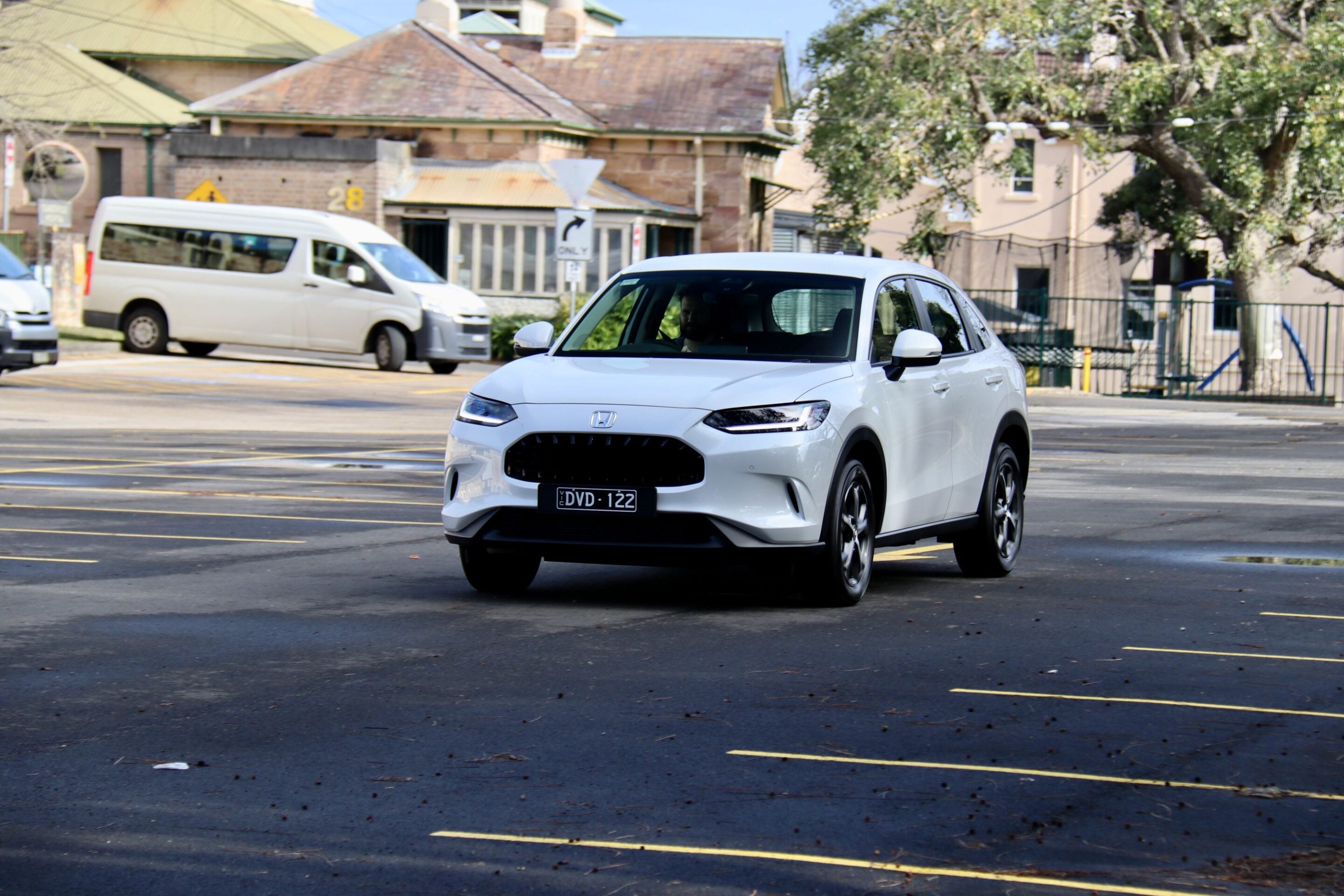 White Honda SUV driving through a paved parking lot with a beige van and brick buildings in the background.