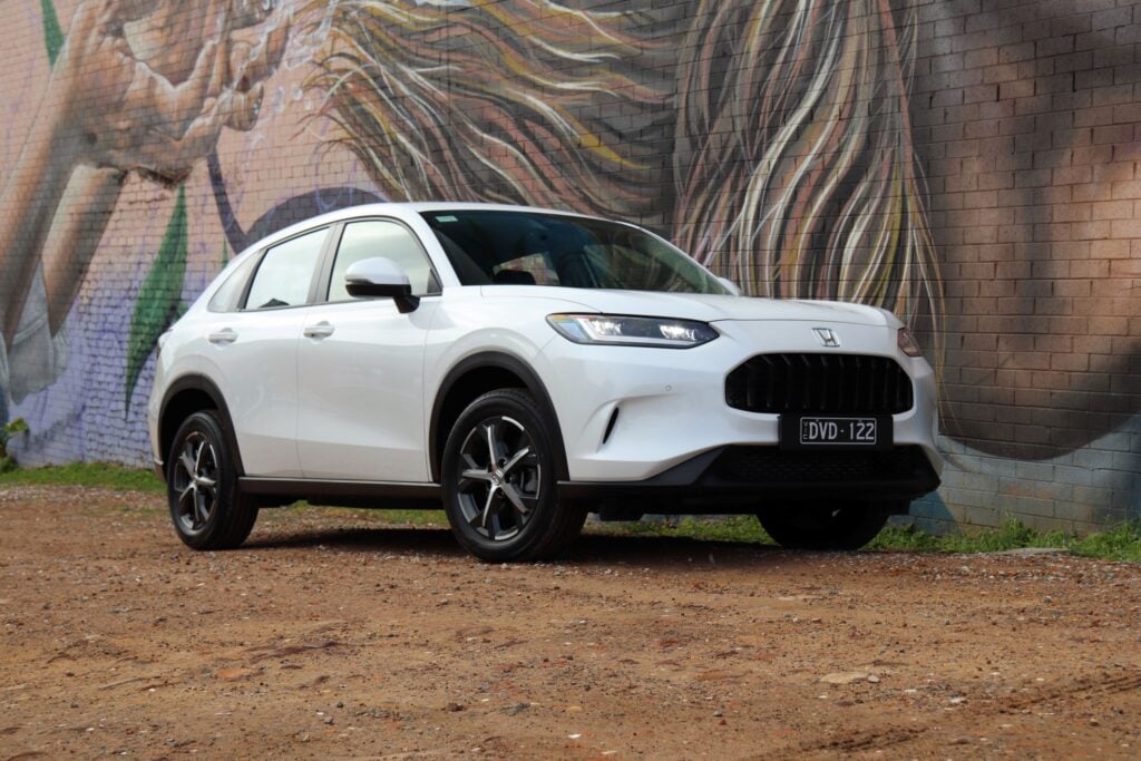 White Honda SUV parked on a dirt lot in front of a large colorful mural painted on a brick wall.