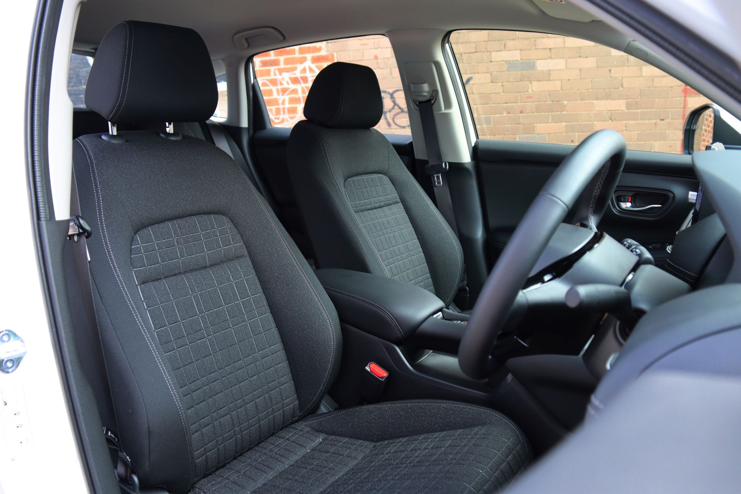 Interior of a car showing front seats with black fabric upholstery and a steering wheel visible on the right side, brick wall outside the window.