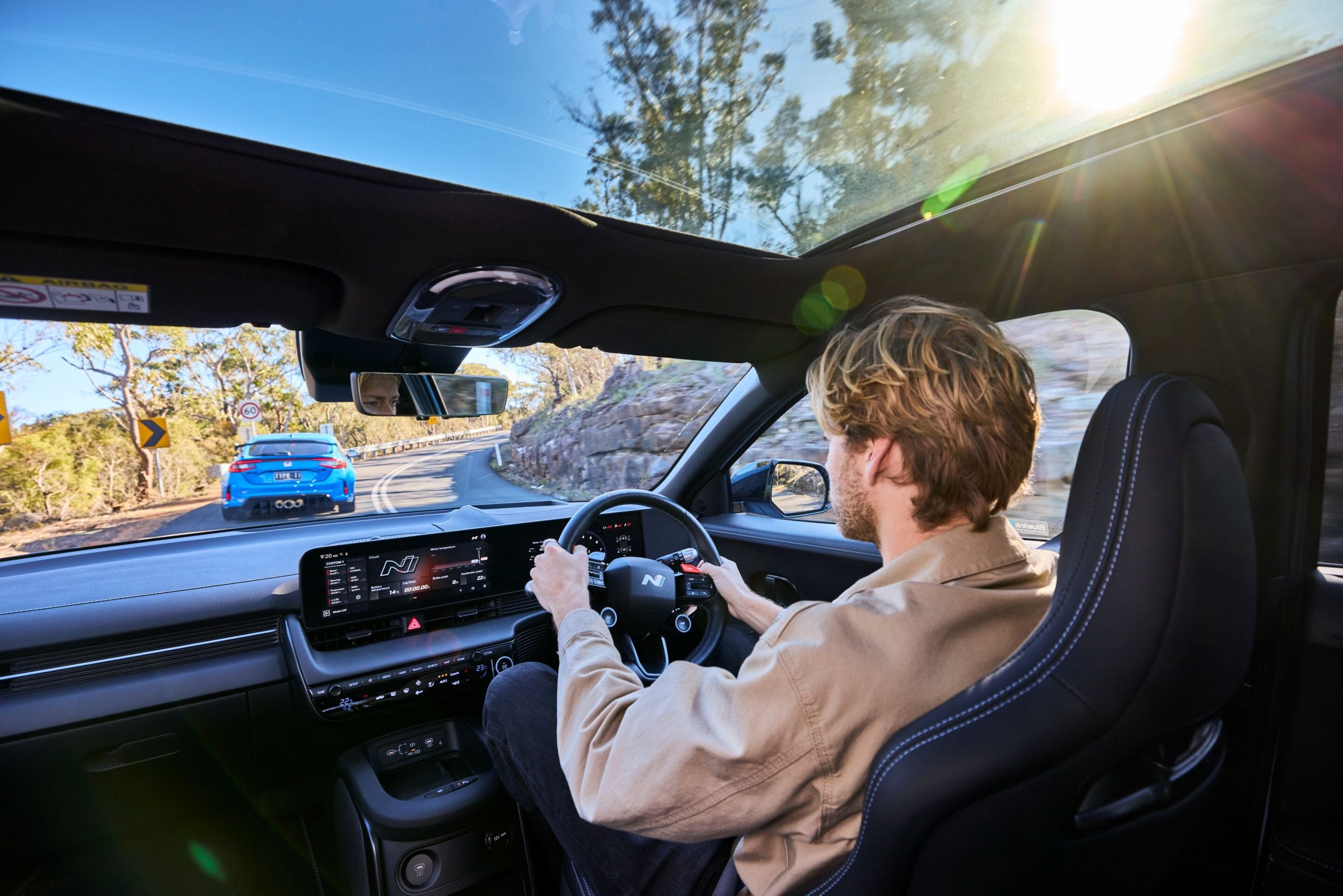 Driver in a beige jacket steers a modern car on a sunny, winding road with a blue car ahead and the dashboard touchscreen visible above the center console.