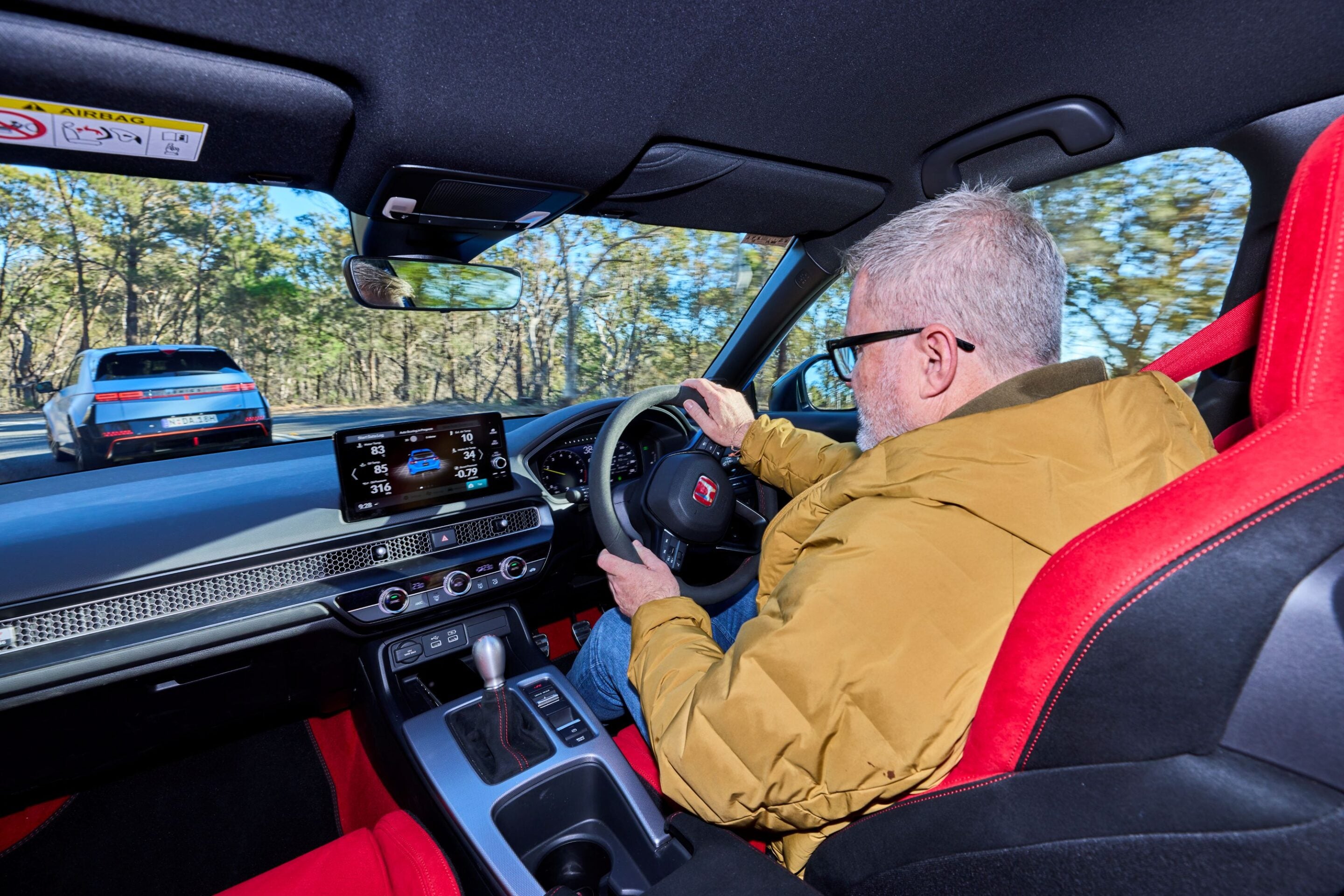Older man with gray hair and glasses driving a car in a tan jacket; digital dashboard screen visible and a blue car is seen on the road behind through the windshield.
